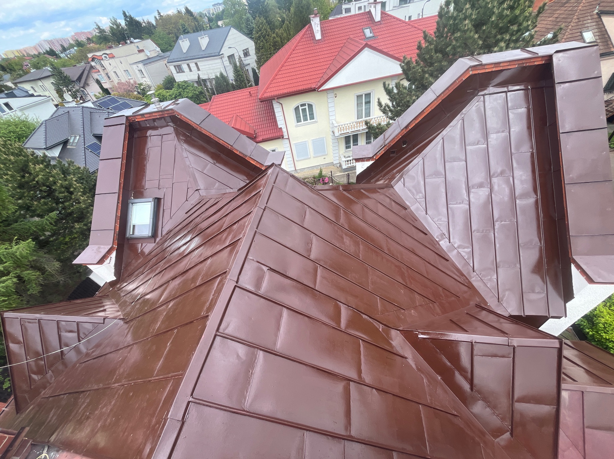 Błyszczący, brązowy dach z blachy trapezowej, ułożony w karo, z widoczną lukarną i fragmentami sąsiednich budynków w tle.