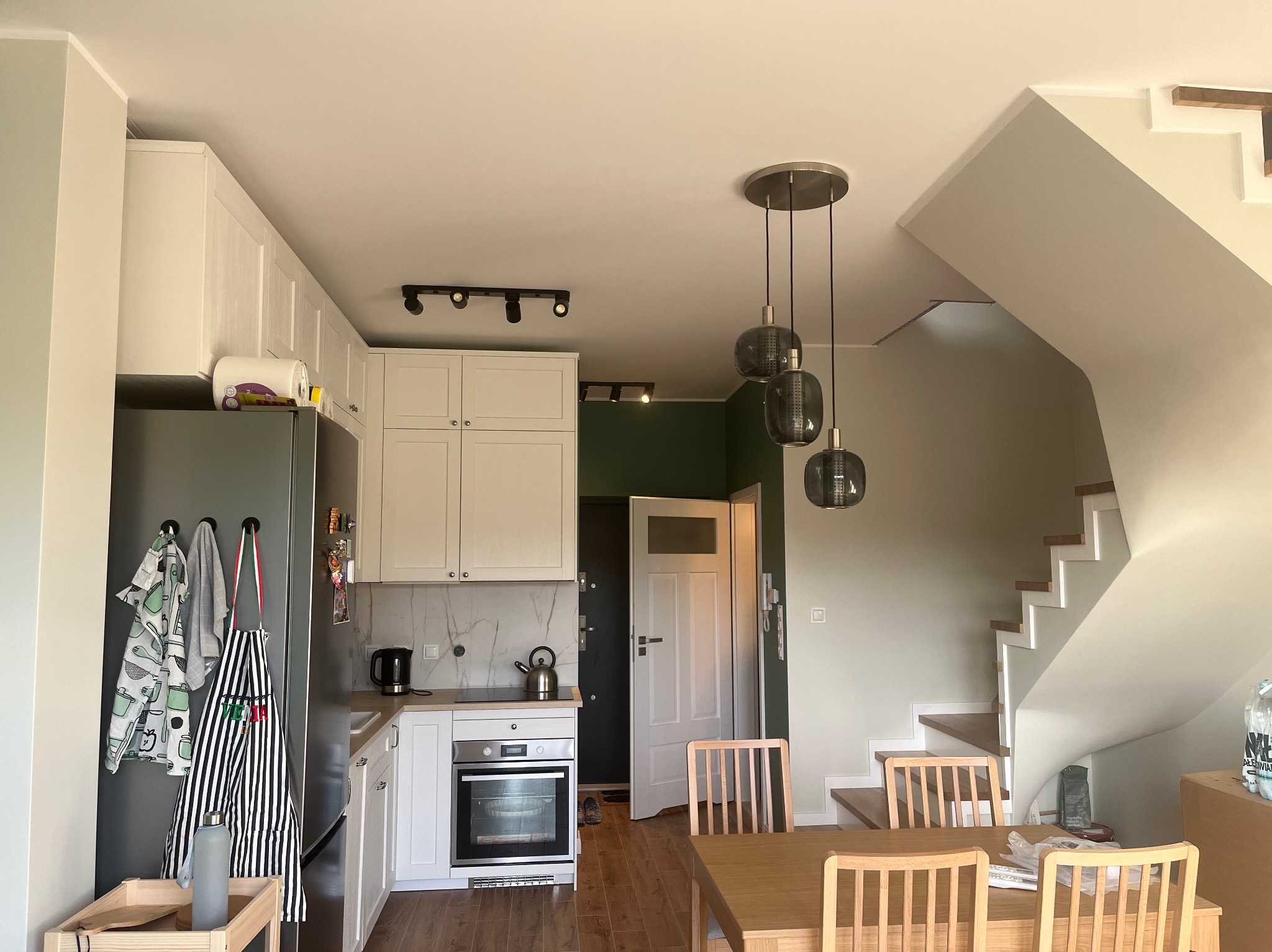 Jasna kuchnia z drewnianym stołem i krzesłami, nowoczesne lampy wiszące, białe meble i schody w tle. Minimalistyczny design, naturalne światło.