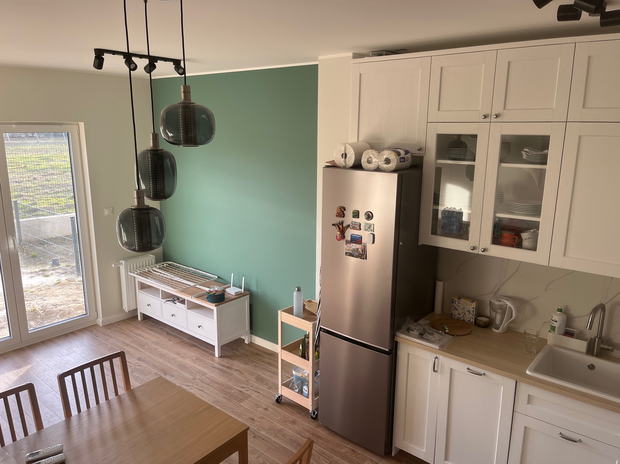 Nowoczesna kuchnia z białymi szafkami, drewnianym blatem i zieloną ścianą. Widoczne są lampy wiszące, stół z krzesłami i lodówka z magnesami.