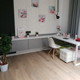 Meblowo Jarosław Warzecha - Pokój z szarą zabudową meblową pod ścianą, białym biurkiem i krzesłem, dekoracjami ściennymi w pastelowych kolorach i drewnianą podłogą.