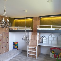 Meblowo Jarosław Warzecha - Pokój dziecięcy z dwoma łóżkami na antresoli, ścianką wspinaczkową, biurkiem z półkami w kształcie domków i lampami wiszącymi w stylu industrialnym.