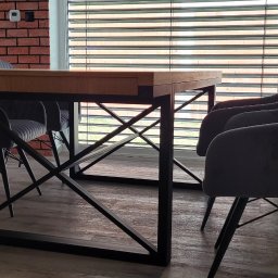Meblowo Jarosław Warzecha - Stół jadalniany z drewnianym blatem i metalowymi nogami w kształcie litery X, otoczony szarymi, tapicerowanymi krzesłami, na tle ceglanej ściany i okna z żaluzjami.