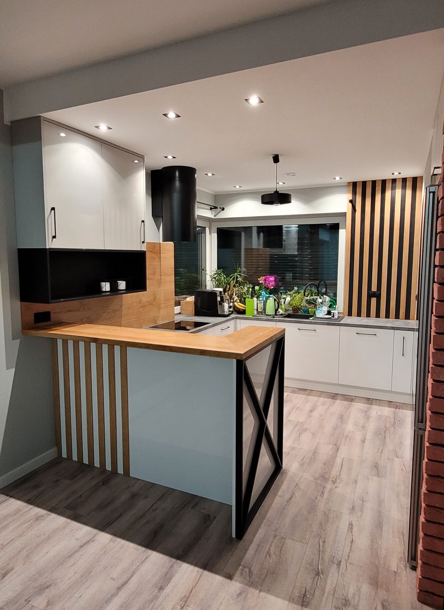 Nowoczesna kuchnia z wyspą, białe szafki, drewniane akcenty i czarny okap cylindryczny, widok na okno z roślinami.