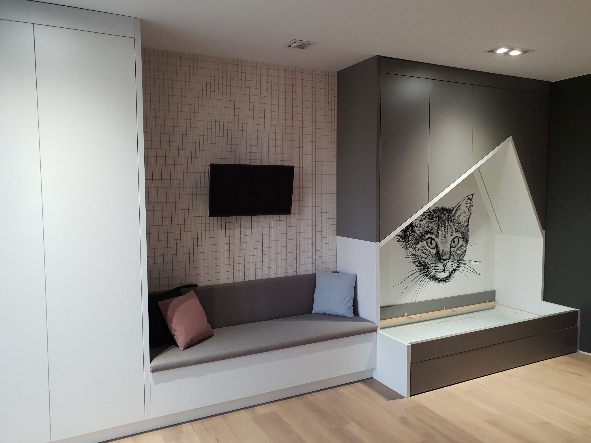 Wnętrze pokoju z zabudową meblową: biała szafa, szara wnęka z siedziskiem z poduszkami, naprzeciwko telewizor na ścianie w kratkę, obok szara szafa z wnęką w kształcie domku z rysunkiem kota.