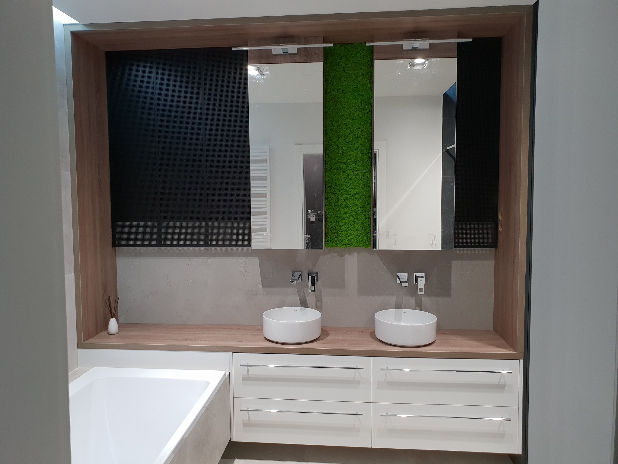 Nowoczesna łazienka z dwiema okrągłymi umywalkami nablatowymi, minimalistyczną armaturą, drewnianym blatem i białymi szufladami, z dekoracyjnym panelem z mchu na ścianie i czarną żaluzją w tle.