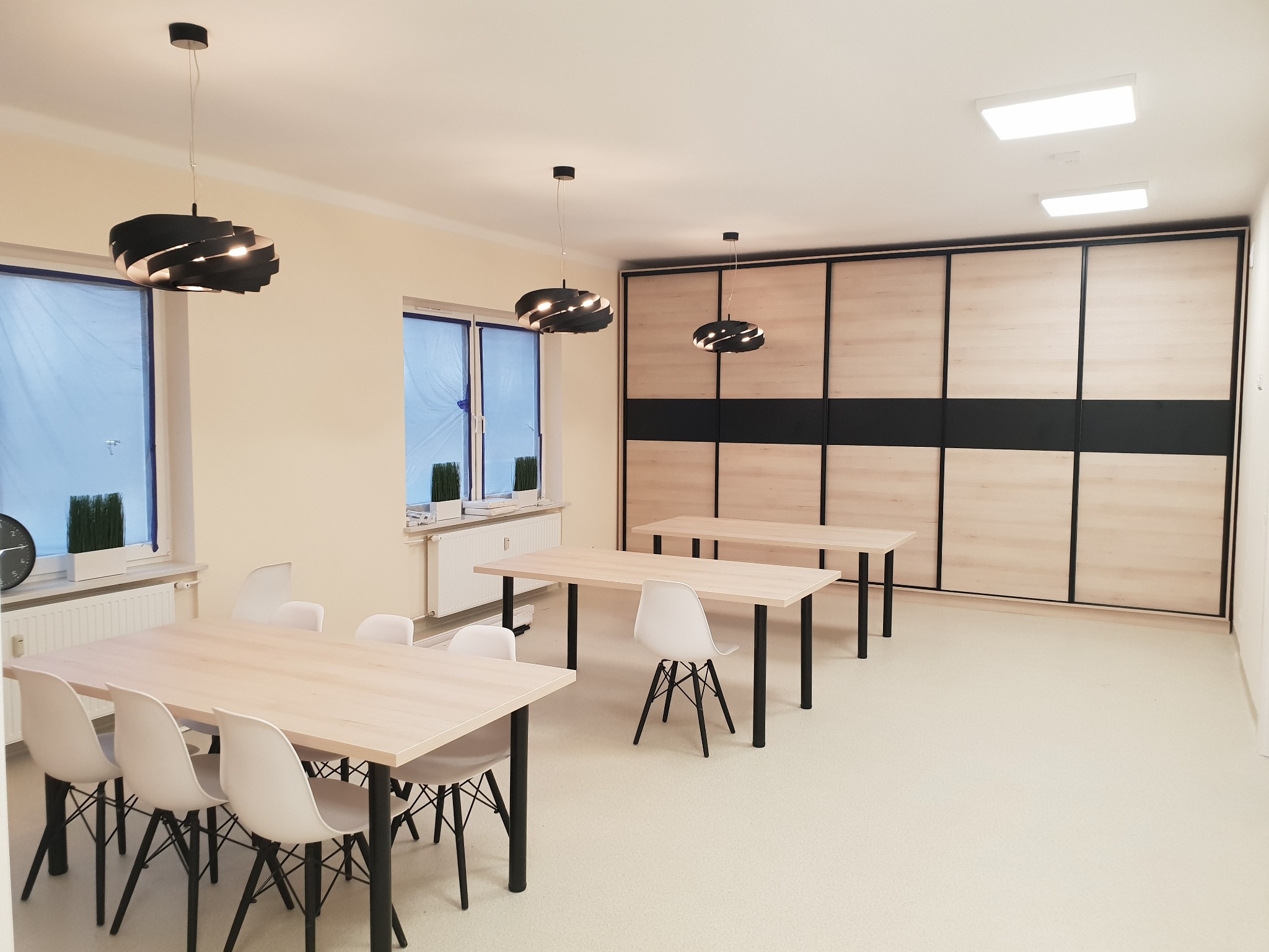 Jasne wnętrze z trzema drewnianymi stołami z czarnymi nogami, białymi krzesłami, dużą przesuwną szafą z drewnianymi panelami i czarnymi obramowaniami oraz trzema nowoczesnymi czarnymi lampami...