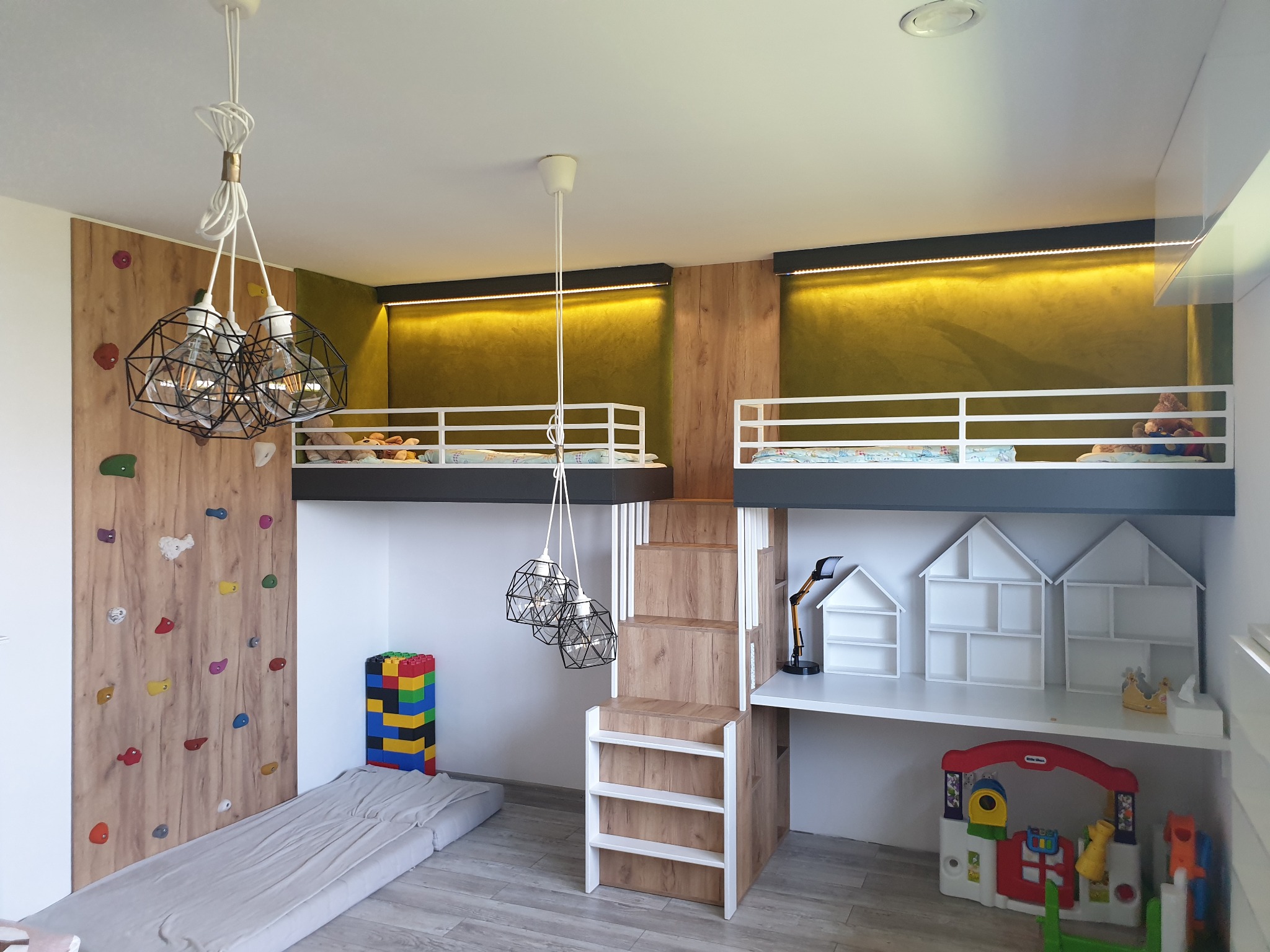Pokój dziecięcy z dwoma łóżkami na antresoli, ścianką wspinaczkową, biurkiem z półkami w kształcie domków i lampami wiszącymi w stylu industrialnym.