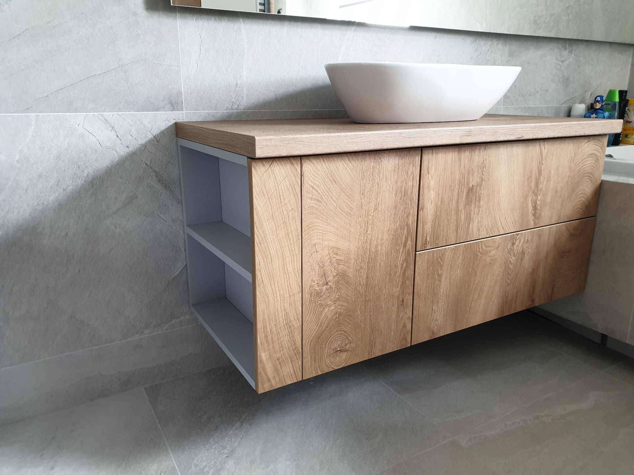 Łazienkowa szafka pod umywalkę nablatową z drewnopodobnymi frontami i otwartymi półkami z boku, szara płytka na ścianie i podłodze, nowoczesny design.