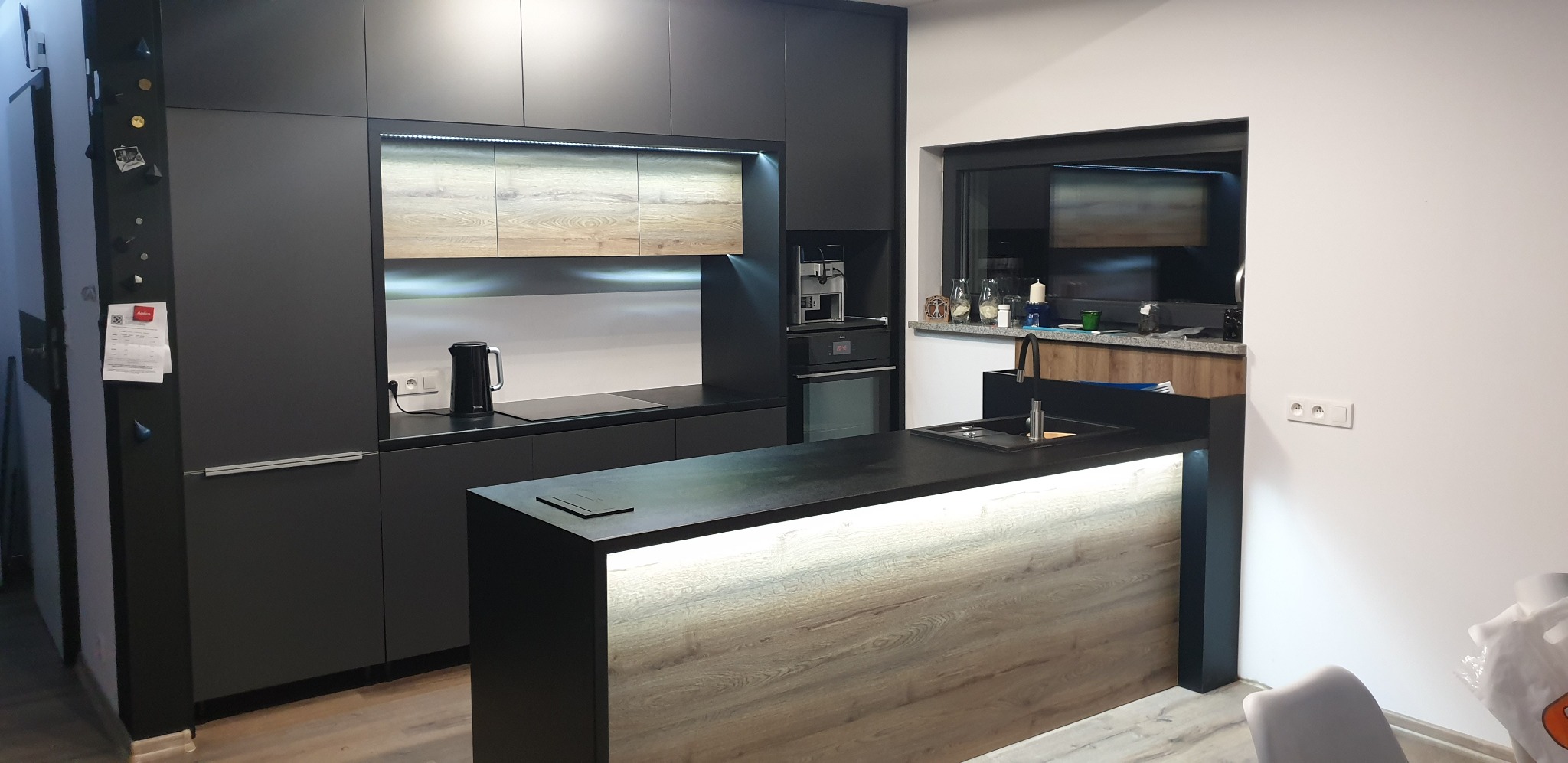 Nowoczesna, czarna kuchnia z wyspą w Gliwicach, z oświetleniem LED pod szafkami i wyspą, widok na ekspres do kawy i okno z dekoracjami.