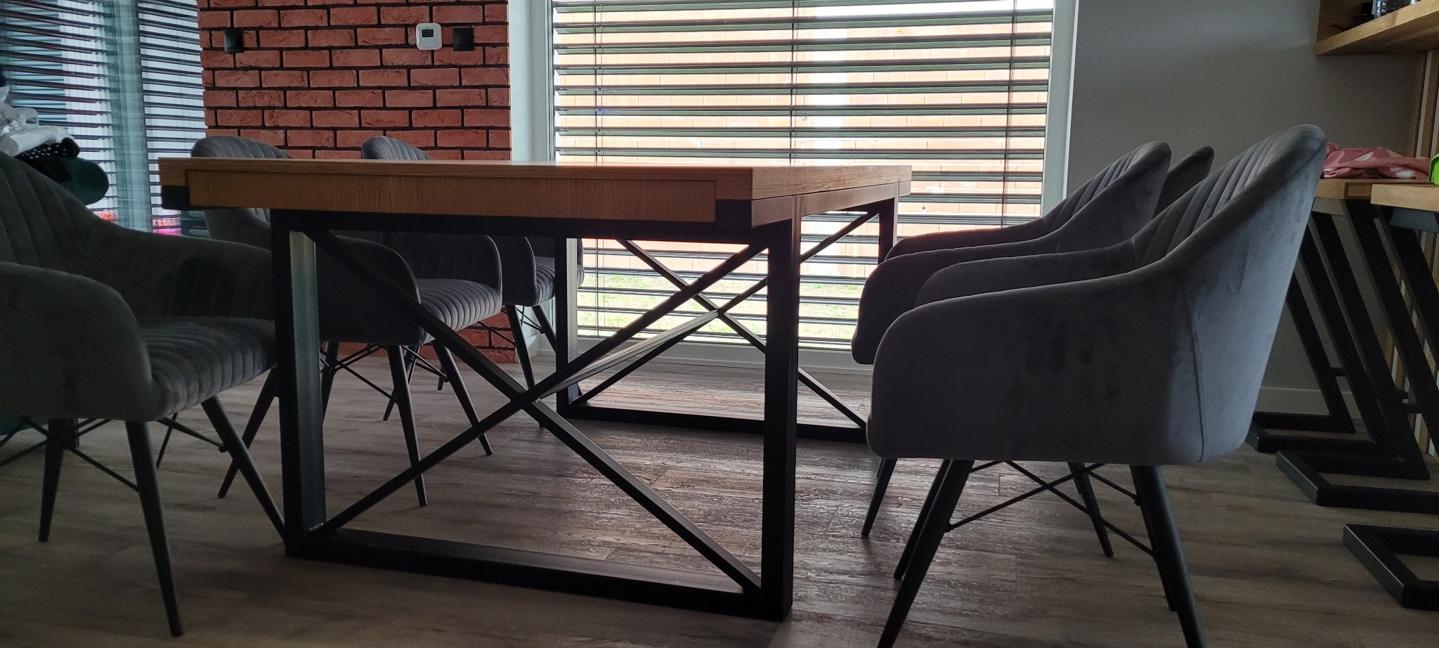 Stół jadalniany z drewnianym blatem i metalowymi nogami w kształcie litery X, otoczony szarymi, tapicerowanymi krzesłami, na tle ceglanej ściany i okna z żaluzjami.