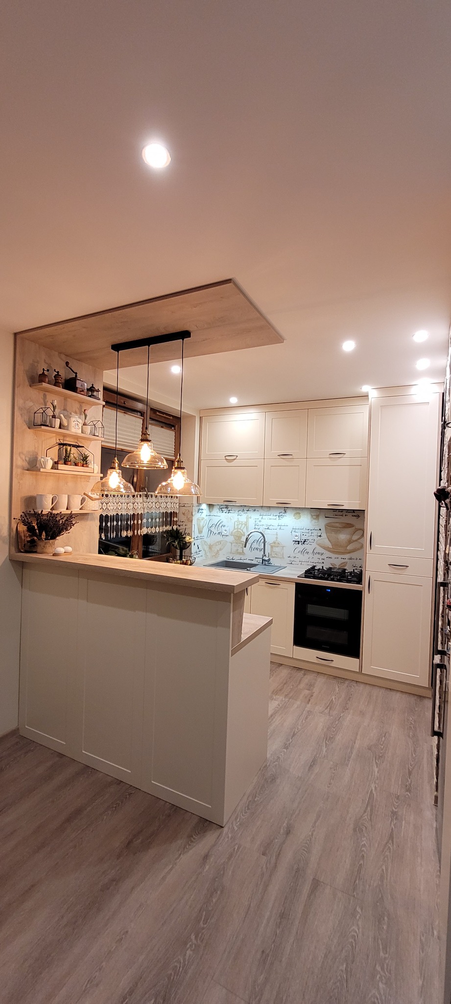 Jasna kuchnia z barkiem, wiszącymi lampami z żarówkami Edisona i dekoracyjną ścianą z motywem kawy.