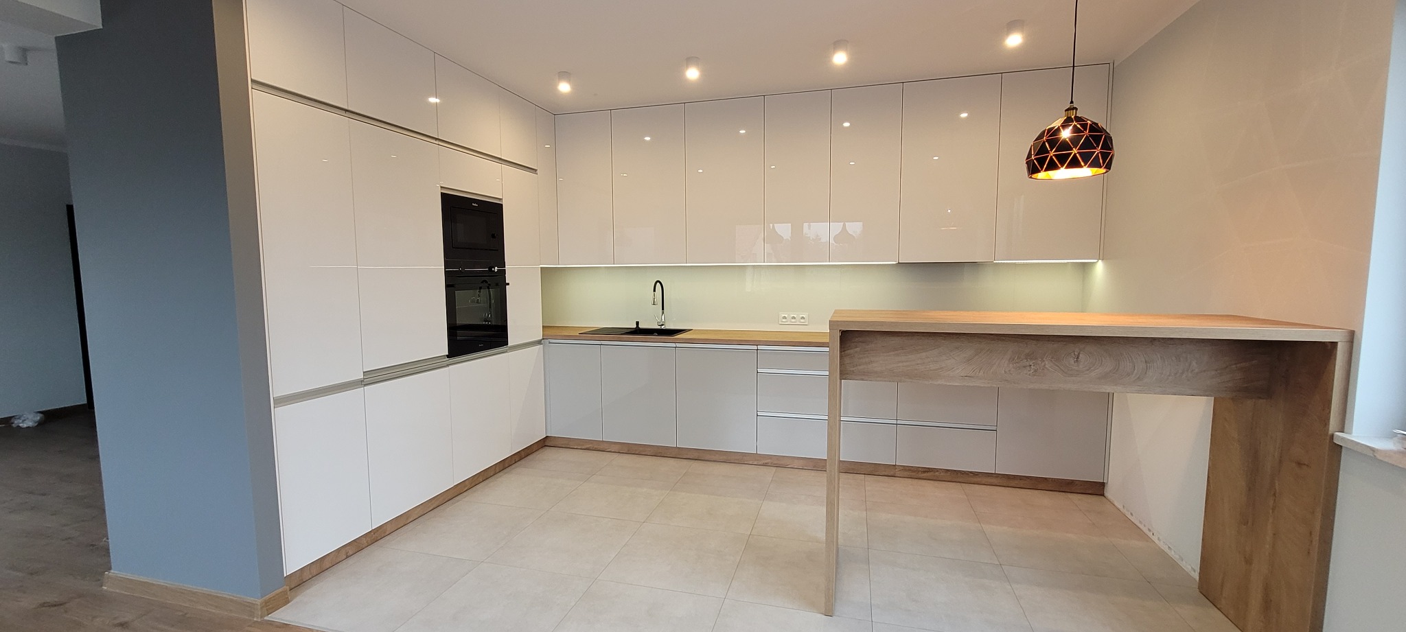 Nowoczesna kuchnia z białymi, lakierowanymi frontami, czarnym zlewem i piekarnikiem w zabudowie, wykończona blatem z jasnego drewna i oświetlona wiszącą lampą z geometrycznym wzorem.
