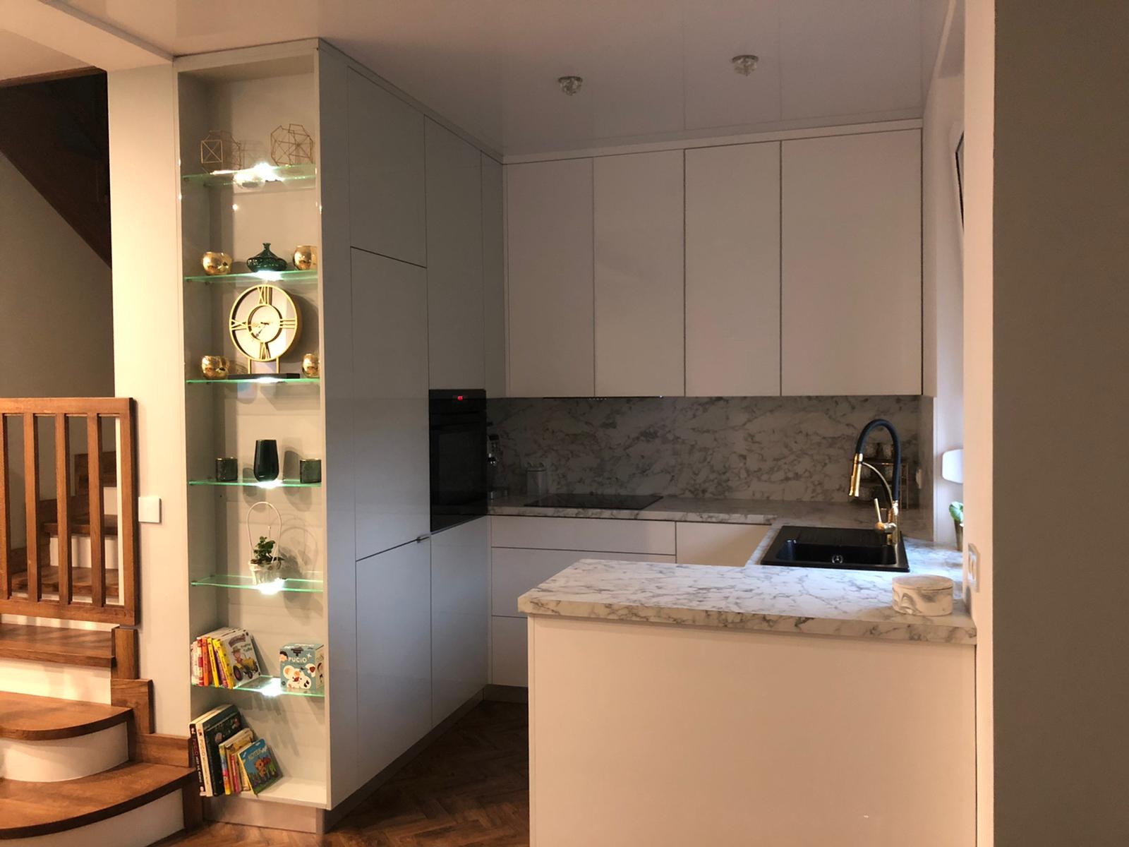 Nowoczesna kuchnia z białymi szafkami, marmurowym blatem i ścianą, zintegrowana z przeszkloną witryną z dekoracjami i książkami, widok na schody z drewnianą balustradą.