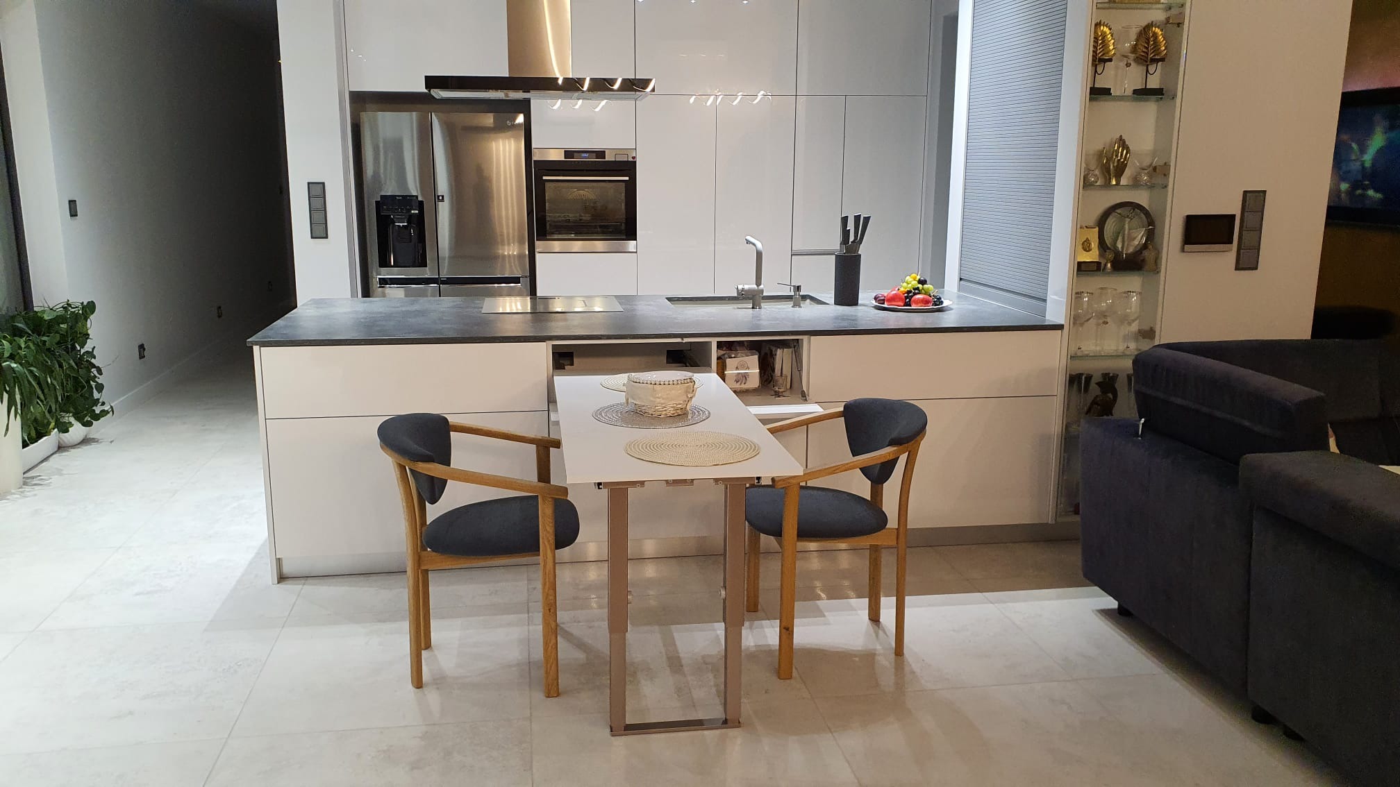 Nowoczesna, biała kuchnia z wysuwanym stołem i dwoma krzesłami, widok na minimalistyczne wnętrze z elementami dekoracyjnymi.