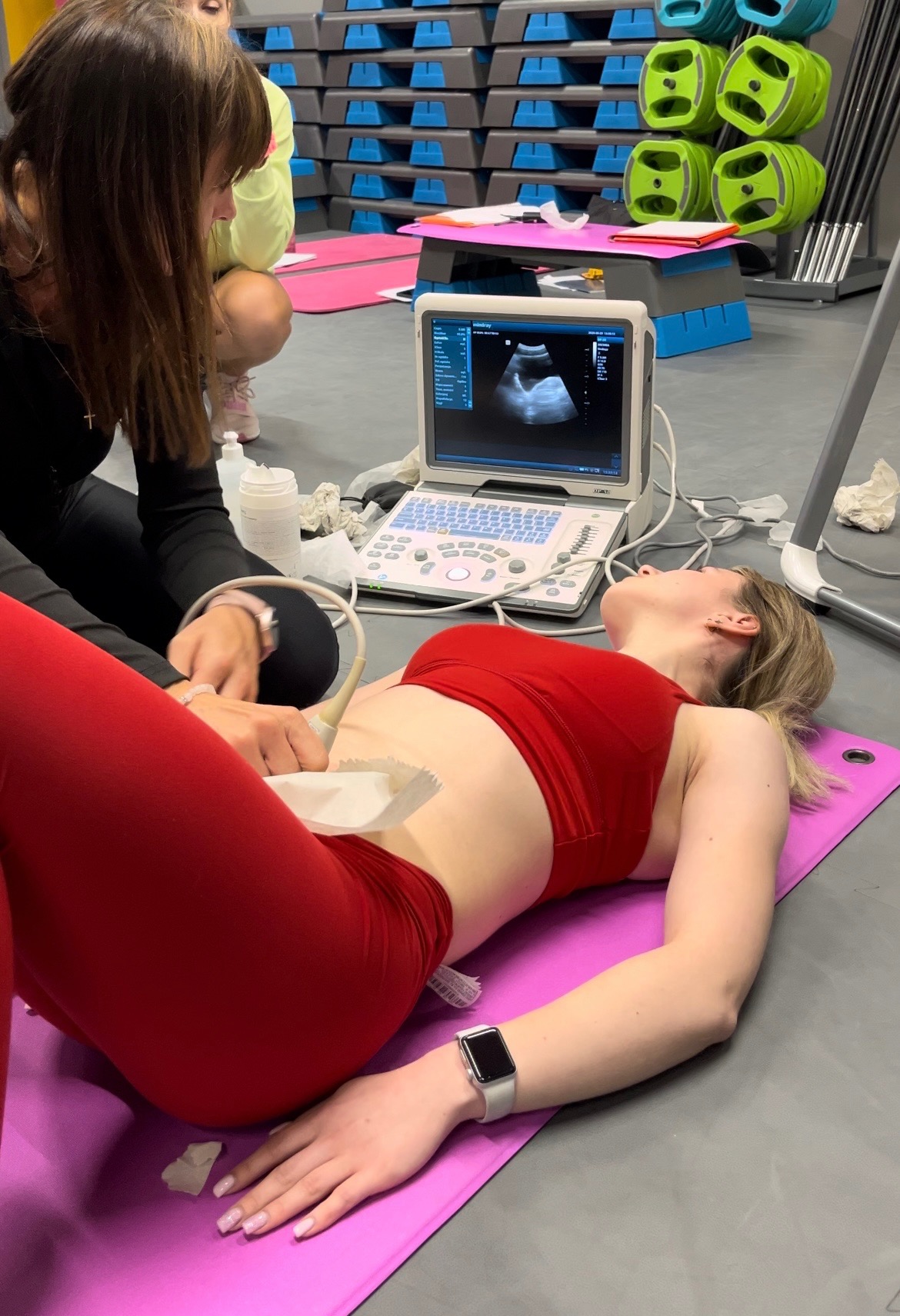 Kobieta leżąca na macie podczas badania USG jamy brzusznej, z widocznym monitorem urządzenia i osobą wykonującą badanie.