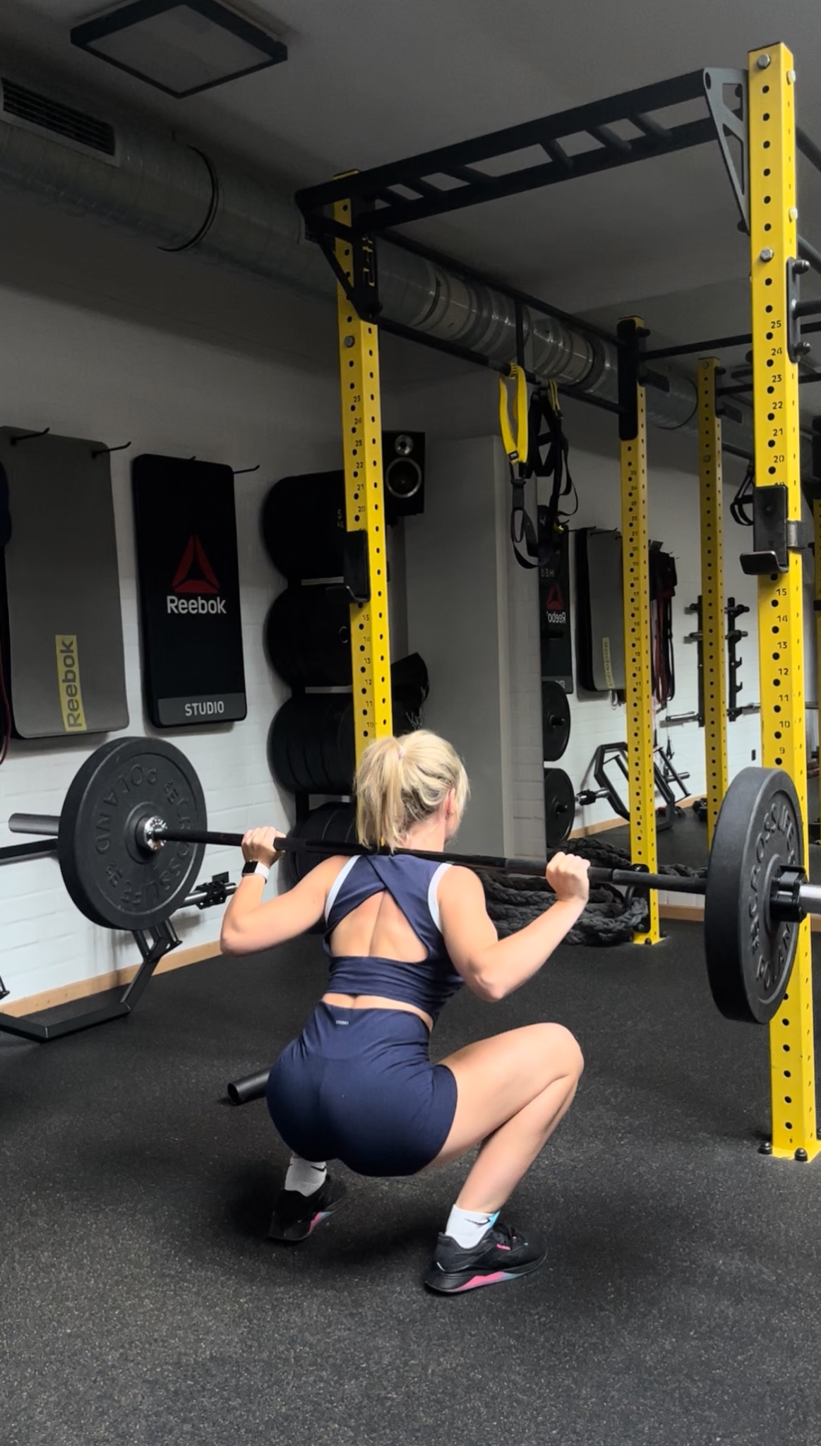 Kobieta w sportowym stroju wykonuje przysiad z obciążeniem w siłowni, widok z tyłu, z żółtymi elementami konstrukcyjnymi i sprzętem Reebok w tle.