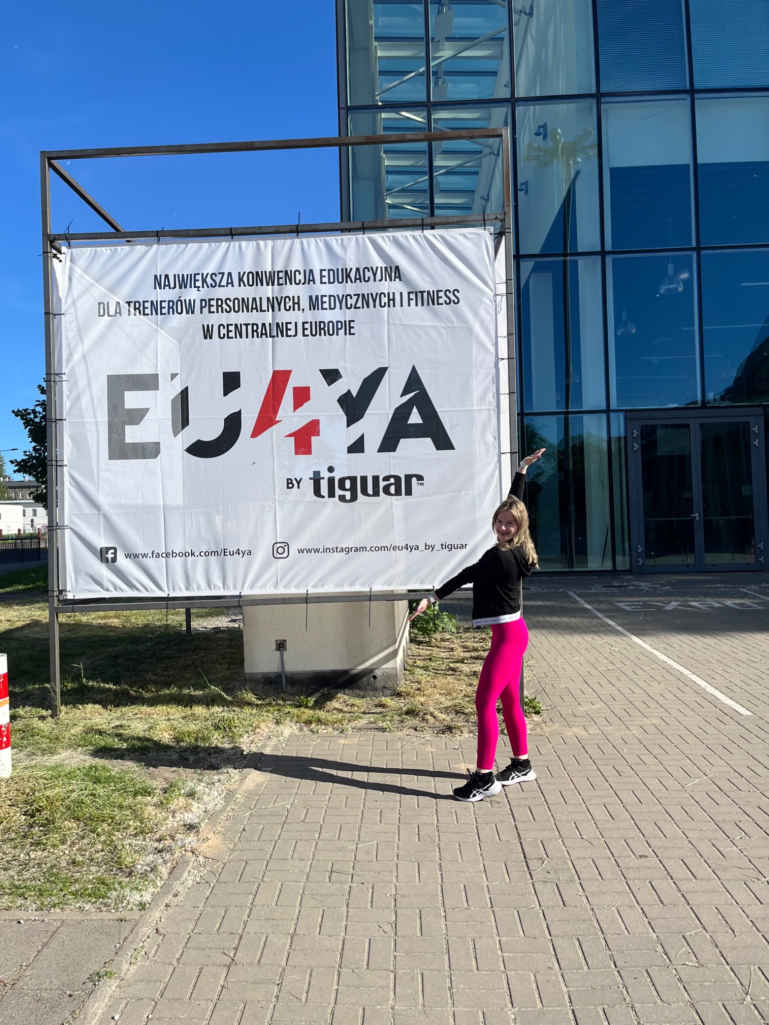 Kobieta w różowych legginsach pozuje przed banerem reklamującym konwencję edukacyjną dla trenerów personalnych, medycznych i fitness o nazwie EU4YA by Tiguar, na tle nowoczesnego budynku...