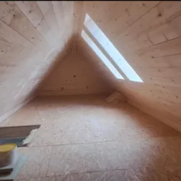 Wnętrze poddasza wykończonego boazerią, z widocznym oknem dachowym w kształcie trójkąta i otworem w podłodze z żółtym wiadrem obok.