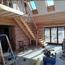 Wojciech SERAFIN FIXER - Wnętrze domu z bali w trakcie budowy, widoczne drewniane schody z linową balustradą, okna dachowe, kompresor i narzędzia budowlane na podłodze, pracownik w tle.