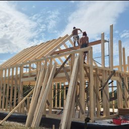 Wojciech SERAFIN FIXER - Konstrukcja szkieletowa domu w trakcie montażu, widoczni dwaj pracownicy na dachu, błękitne niebo z chmurami w tle.