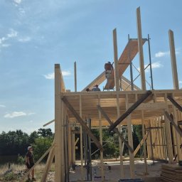 Wojciech SERAFIN FIXER - Wznoszenie konstrukcji drewnianej domu w słoneczny dzień, widoczni dwaj pracownicy na rusztowaniu i trzeci na ziemi, rozrzucone materiały budowlane w otoczeniu.