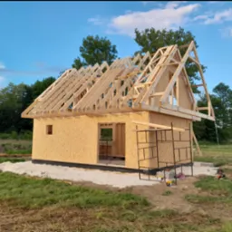 Wznoszenie konstrukcji drewnianego domu z płyt OSB, widoczne rusztowanie i elementy więźby dachowej na tle zieleni i błękitnego nieba.