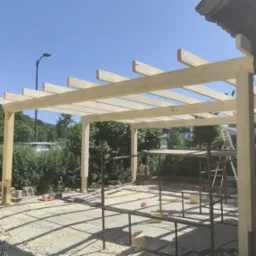 Drewniana konstrukcja z belek, prawdopodobnie zadaszenie tarasu, w trakcie budowy. Widoczne rusztowania i narzędzia. Tło: zieleń i błękitne niebo.