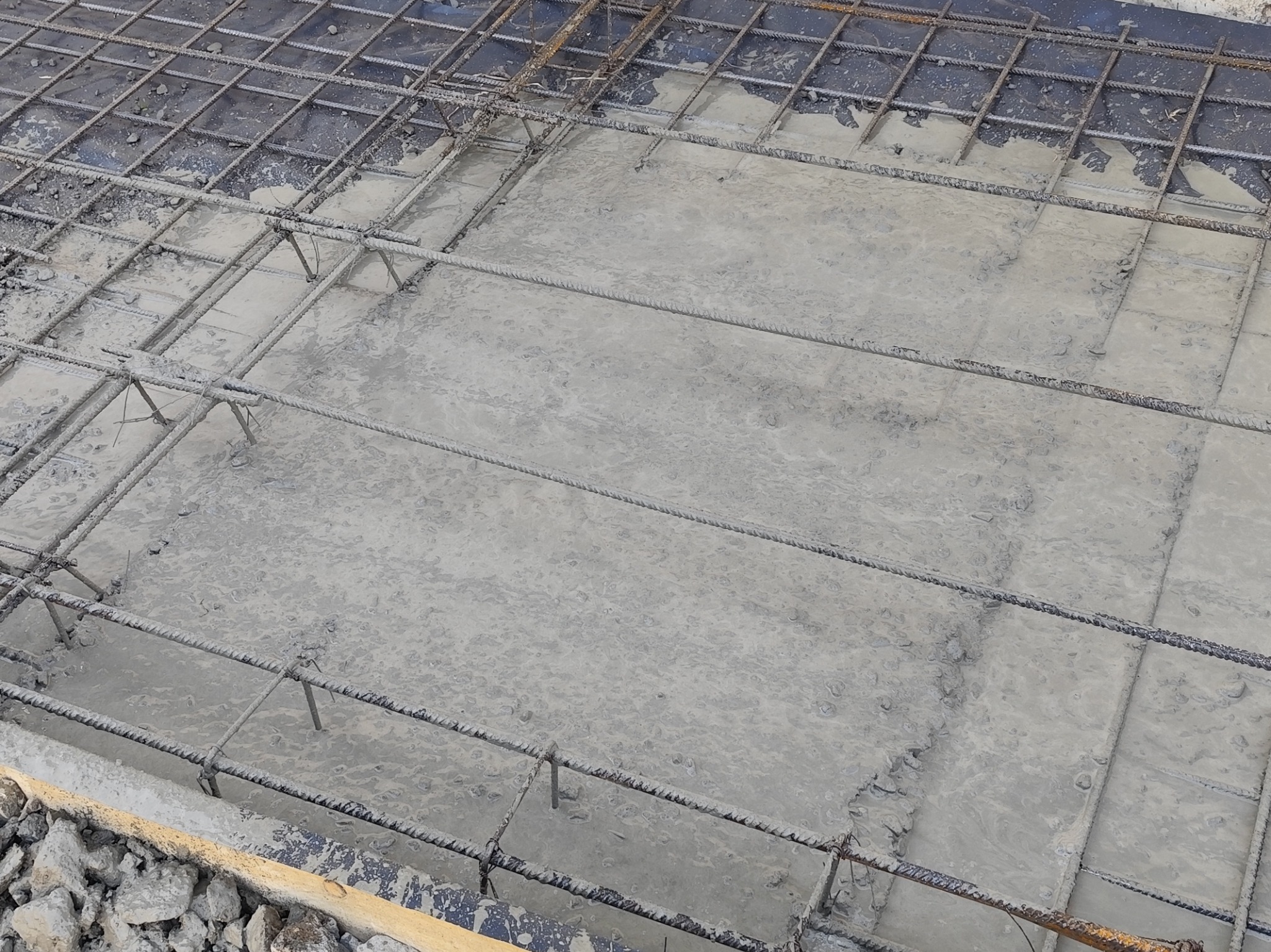 Zalewanie betonem zbrojonej siatki stalowej na budowie, widoczne fragmenty szalunku i kamienie w tle.