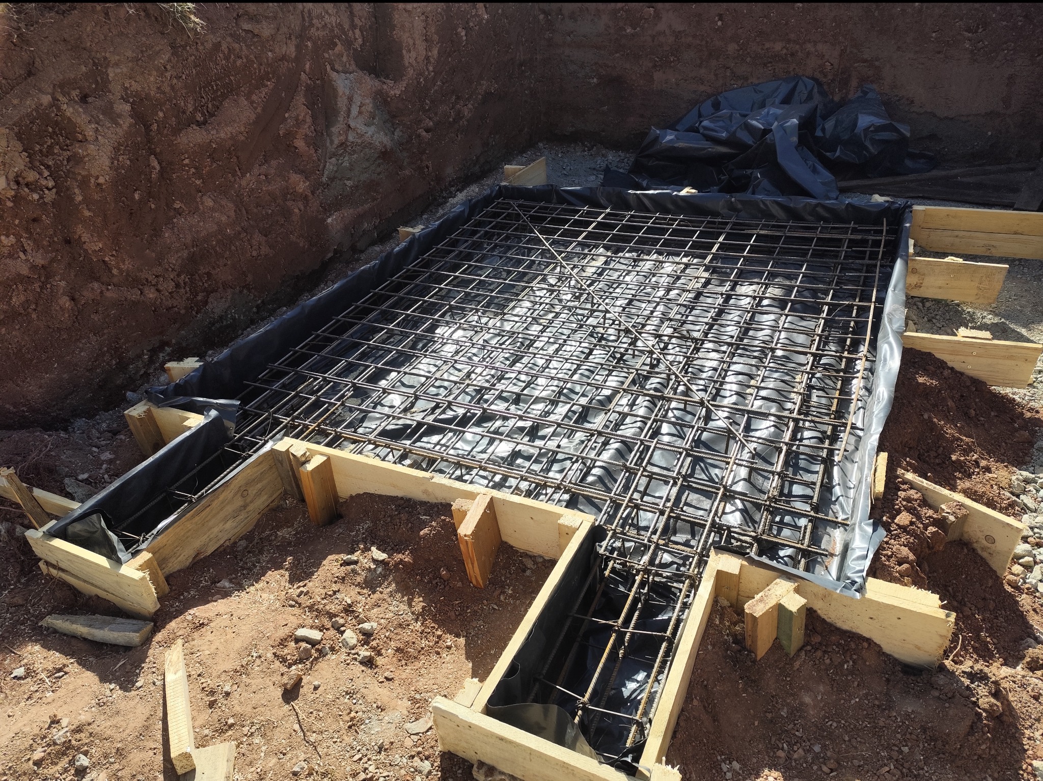 Zbrojenie fundamentów z siatki stalowej i deskowania drewnianego, zabezpieczone folią hydroizolacyjną, w wykopie ziemnym przygotowanym pod wylewkę.