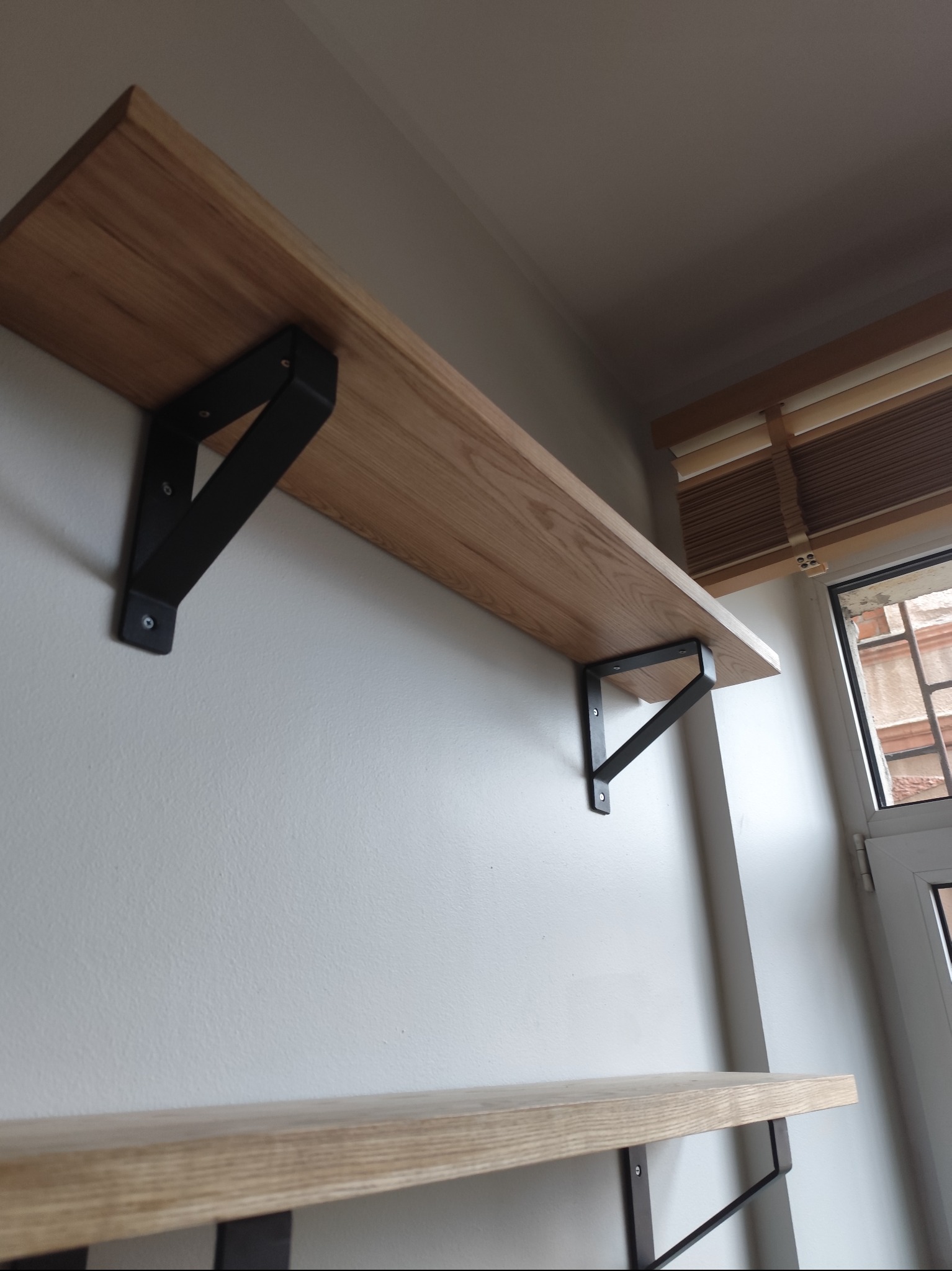 Półka z jasnego drewna zamontowana na ścianie za pomocą czarnych, metalowych wsporników, widoczne okno z żaluzjami w tle.