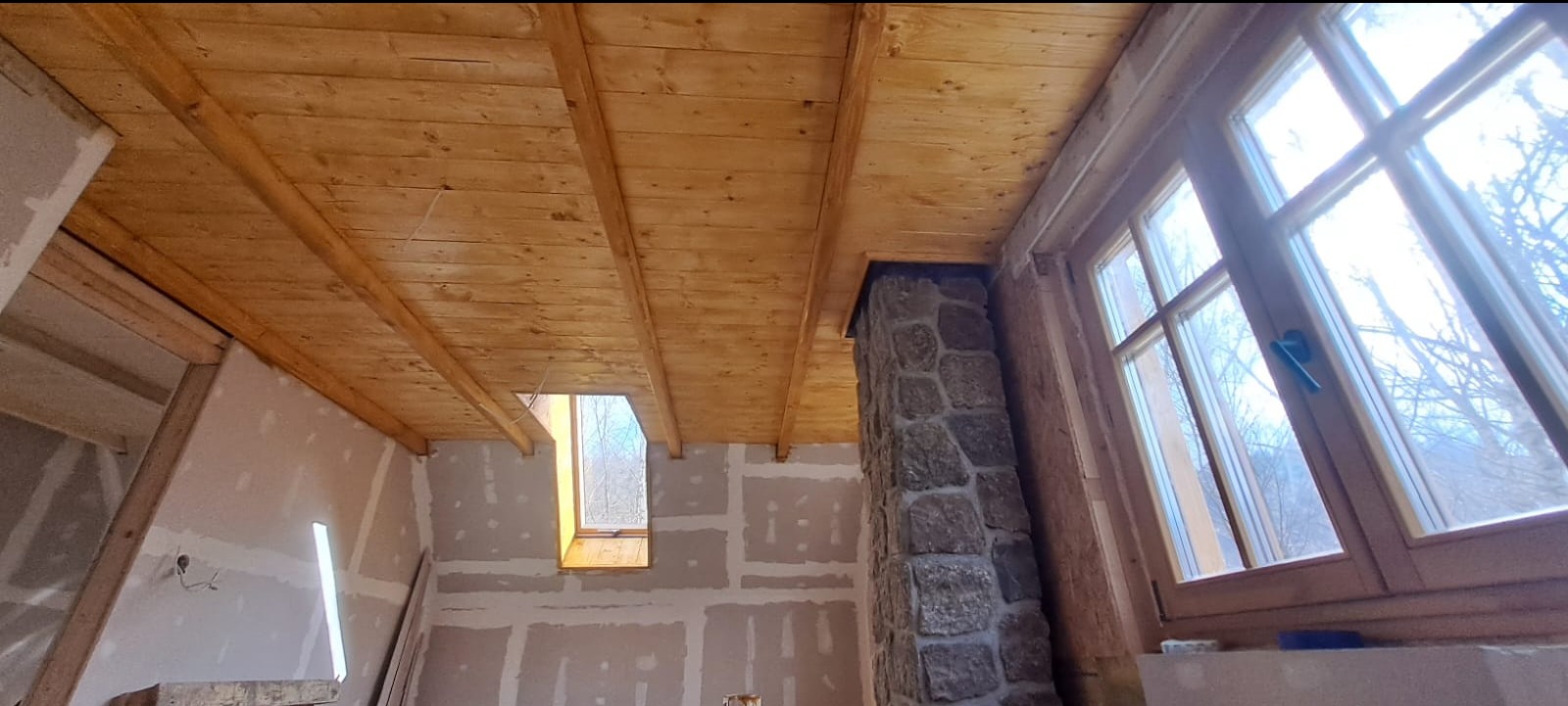 Wnętrze surowego domu w trakcie budowy: drewniany strop z widocznymi belkami, ściany wyłożone płytami kartonowo-gipsowymi, kamienny komin oraz okno z widokiem na drzewa.