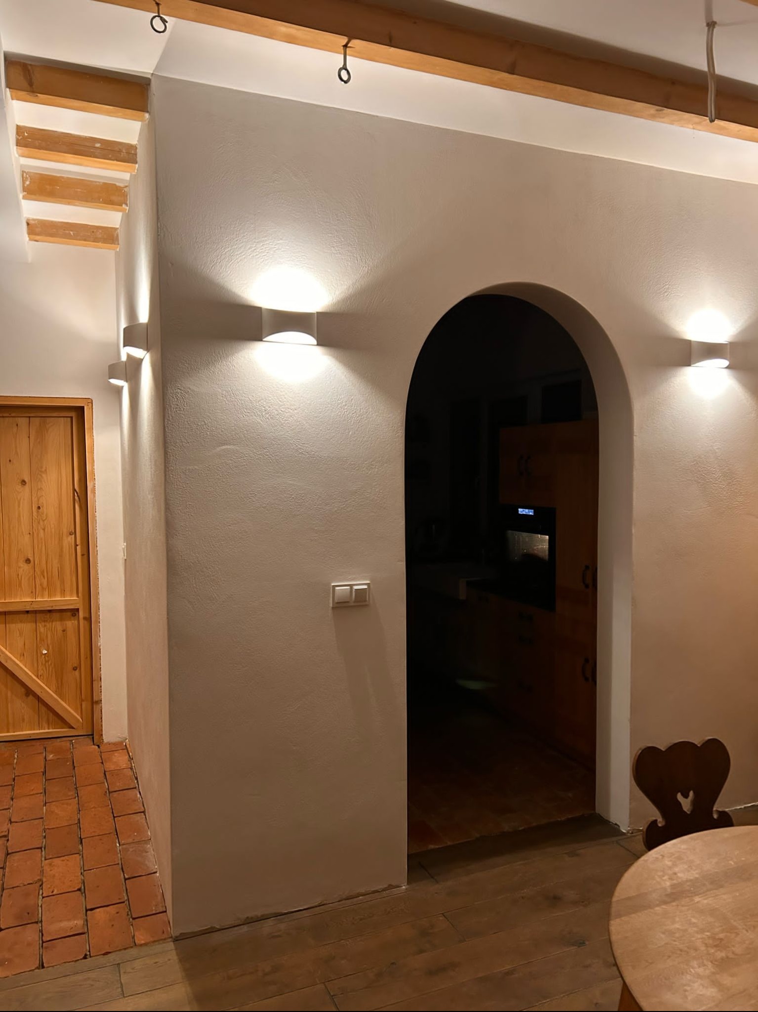 Wnętrze domu z łukowatym przejściem do kuchni, oświetlone kinkietami na ścianach, z widocznym drewnianym sufitem i ceglaną podłogą przy drzwiach.