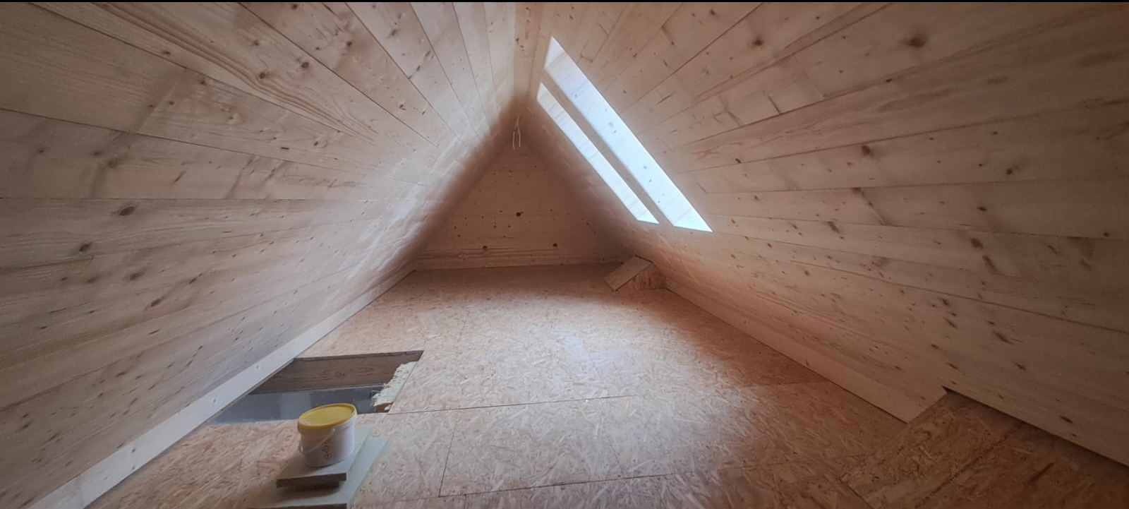 Wnętrze poddasza wykończonego boazerią, z widocznym oknem dachowym w kształcie trójkąta i otworem w podłodze z żółtym wiadrem obok.