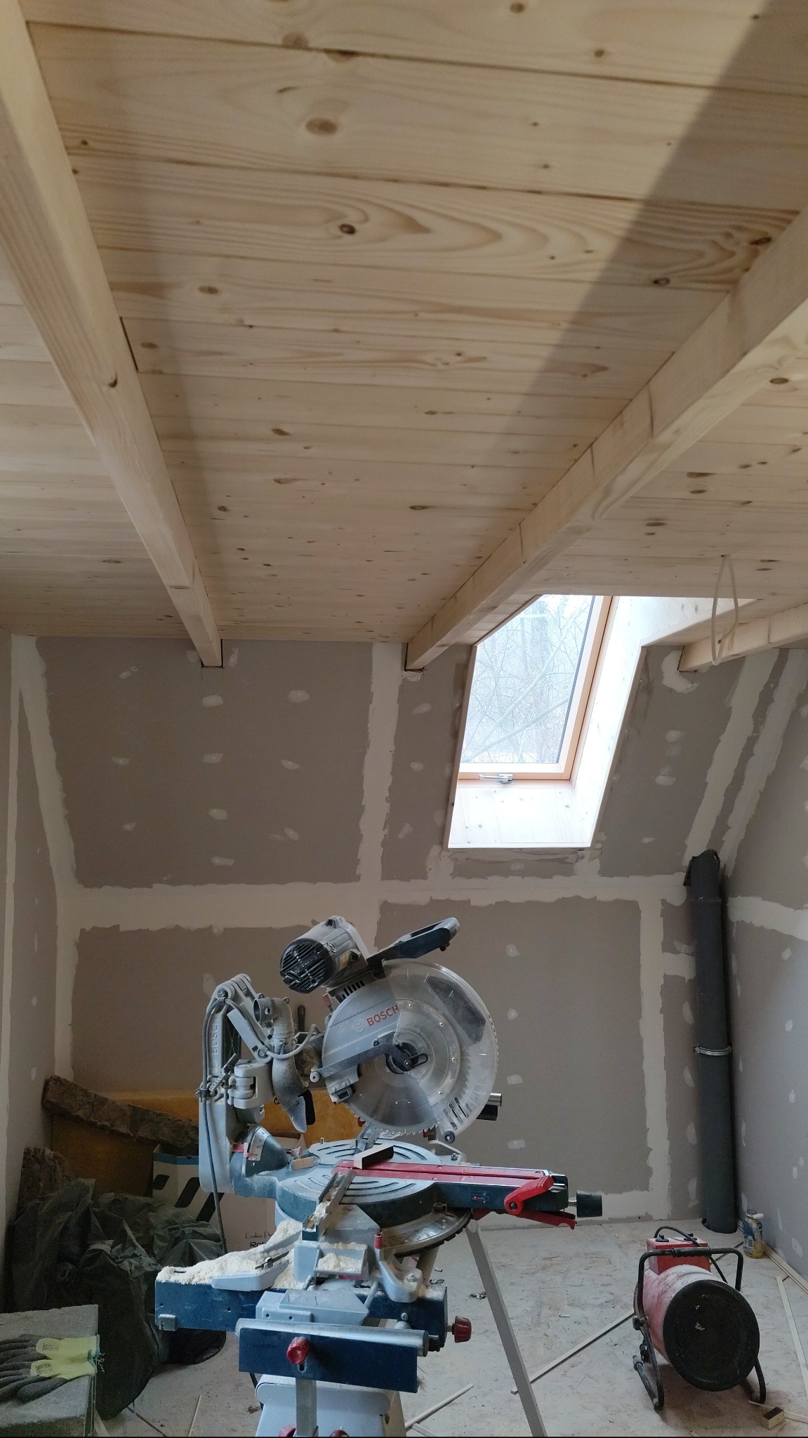 Poddasze w trakcie wykańczania: drewniana boazeria na suficie, okno dachowe, ściany z płyt gipsowo-kartonowych przed szpachlowaniem, piła ukośnica Bosch na pierwszym planie.