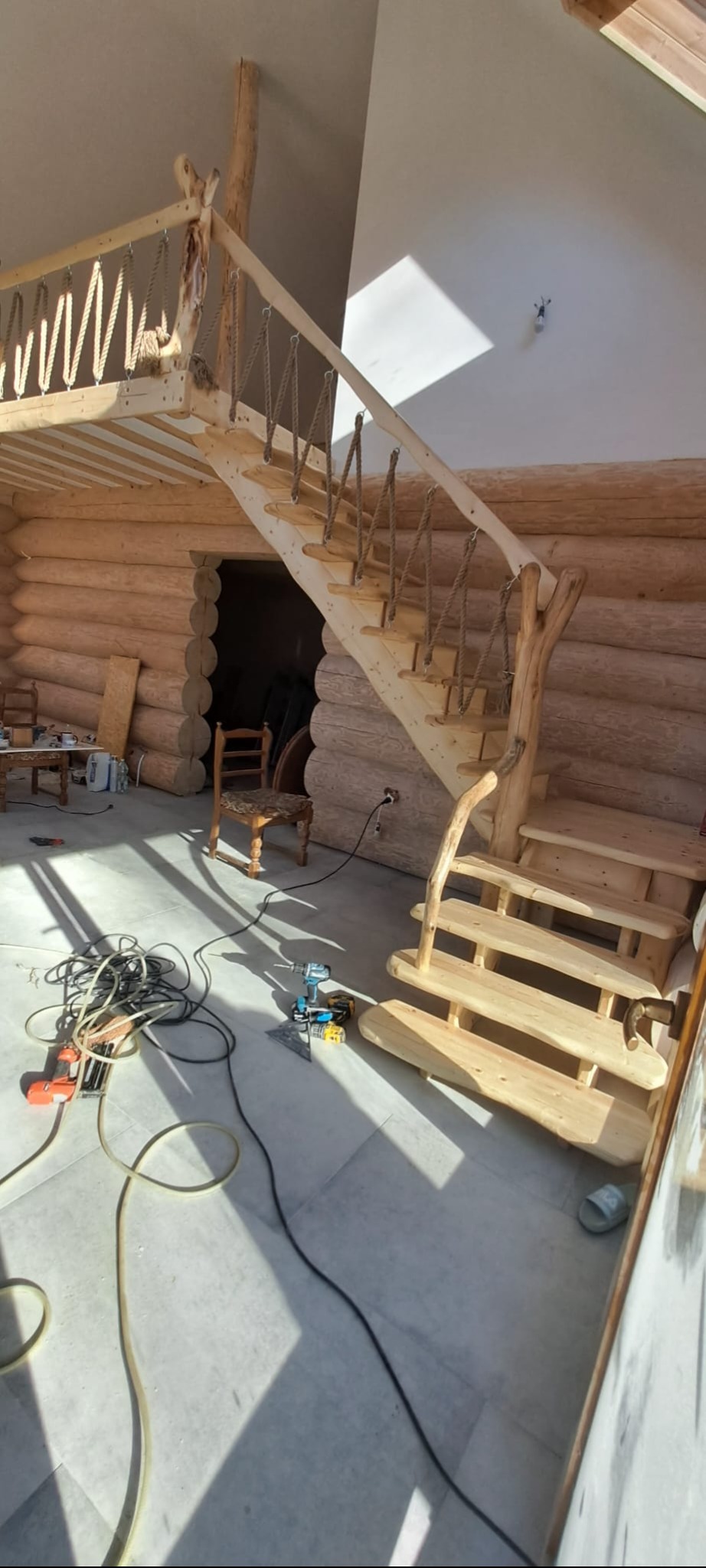 Wnętrze domu z bali z drewnianymi schodami o nietypowej konstrukcji, zabezpieczonymi linami zamiast balustrady, w tle widoczne narzędzia budowlane i krzesło.