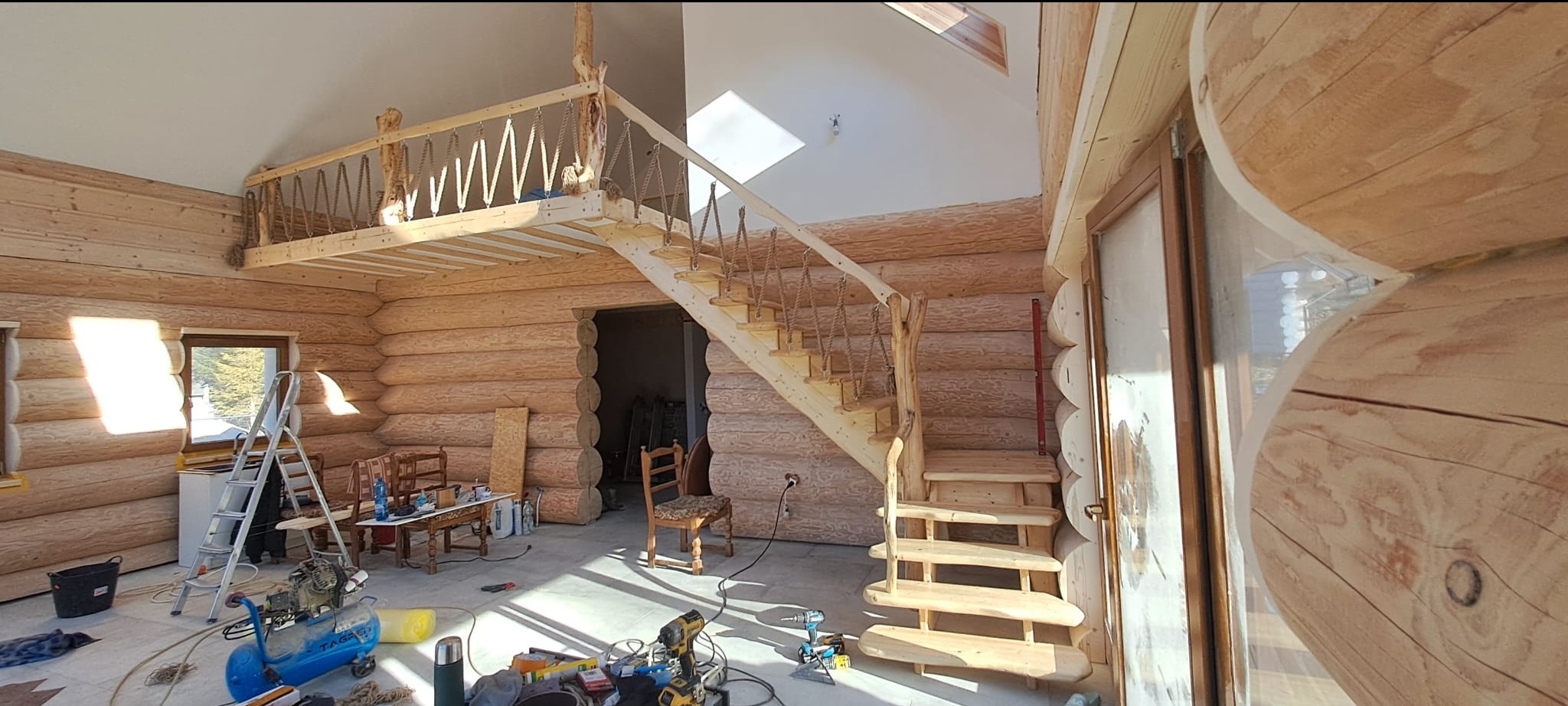 Wnętrze domu z bali w Stroniu Śląskim w trakcie wykańczania, z widocznymi drewnianymi schodami z linowymi poręczami, antresolą i narzędziami na podłodze.