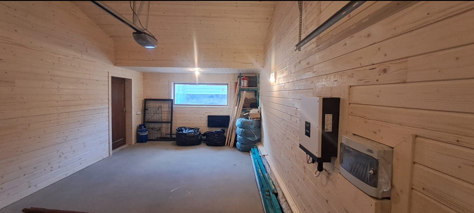 Wnętrze garażu wykończonego jasnym drewnem, z widocznym mechanizmem bramy garażowej pod sufitem, oknem w głębi i zainstalowanym inwerterem fotowoltaicznym na ścianie.