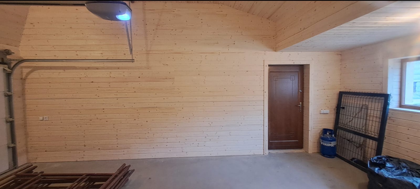 Wnętrze garażu wykończonego boazerią z jasnego drewna, widoczna brama garażowa z automatyką, brązowe drzwi wejściowe, okno, butla gazowa i metalowa krata.