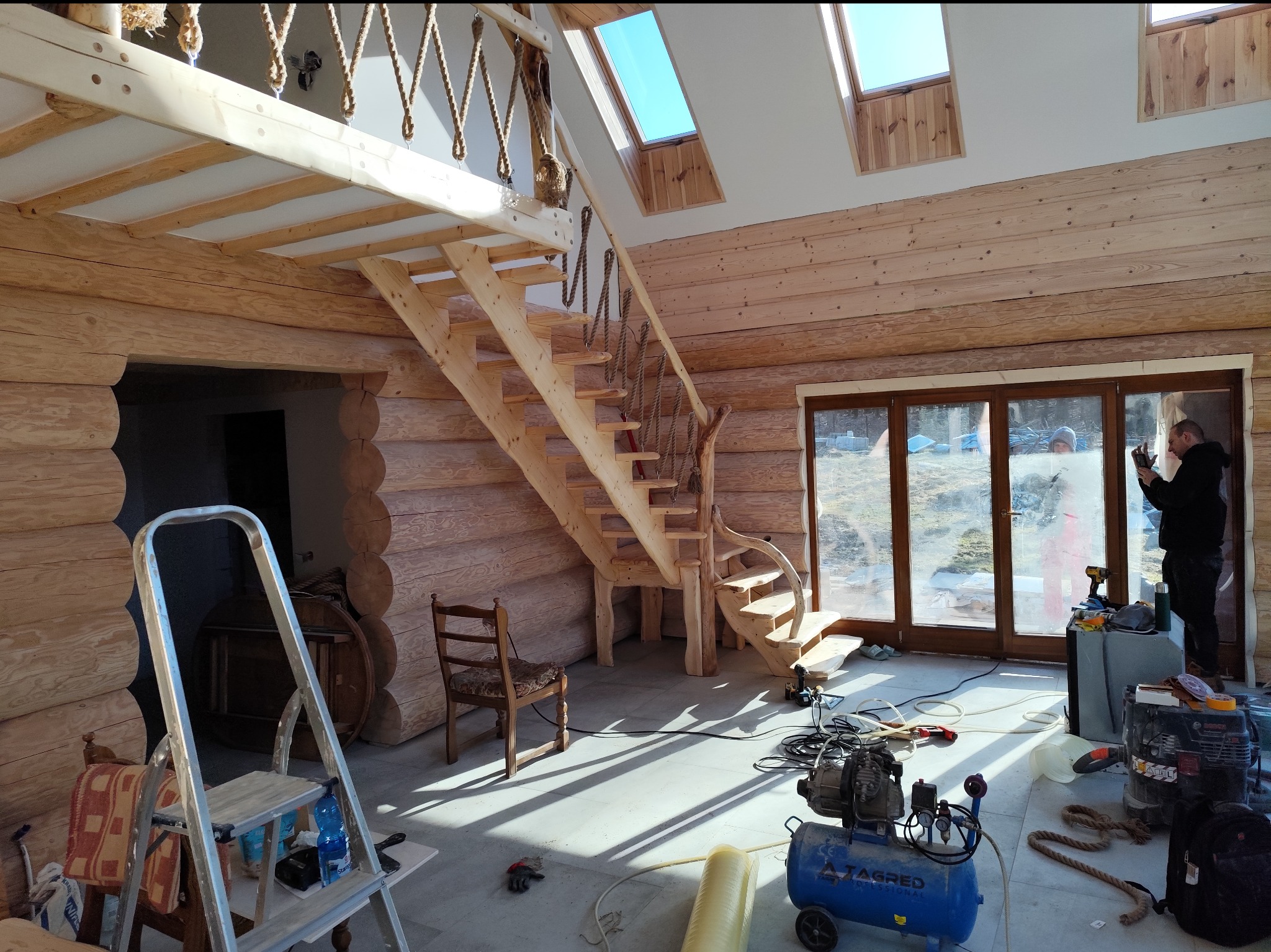 Wnętrze domu z bali w trakcie budowy, widoczne drewniane schody z linową balustradą, okna dachowe, kompresor i narzędzia budowlane na podłodze, pracownik w tle.