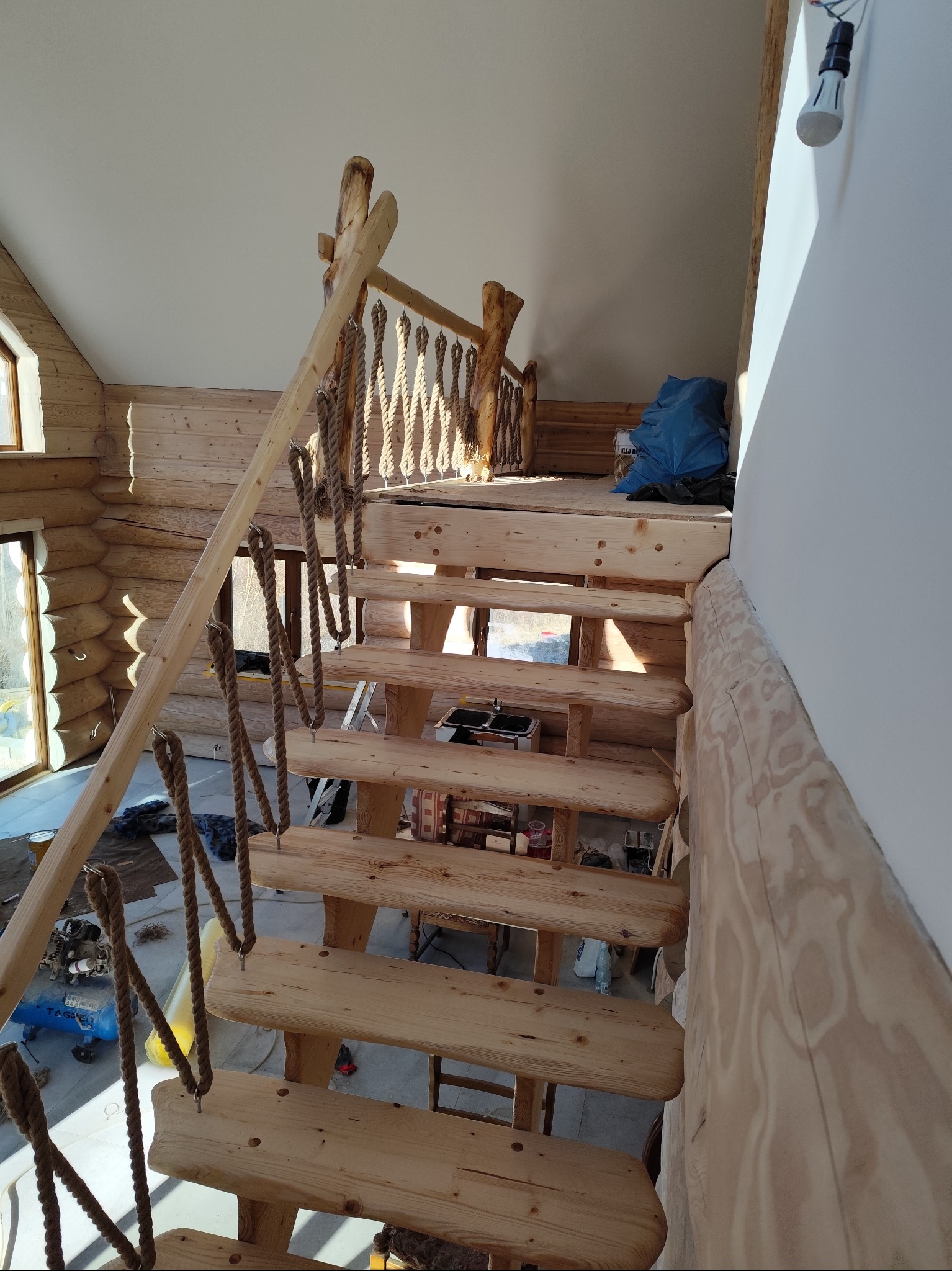 Wnętrze domu z bali z surowymi, drewnianymi schodami z linowymi poręczami, widok z góry na częściowo wykończone schody i narzędzia na podeście.