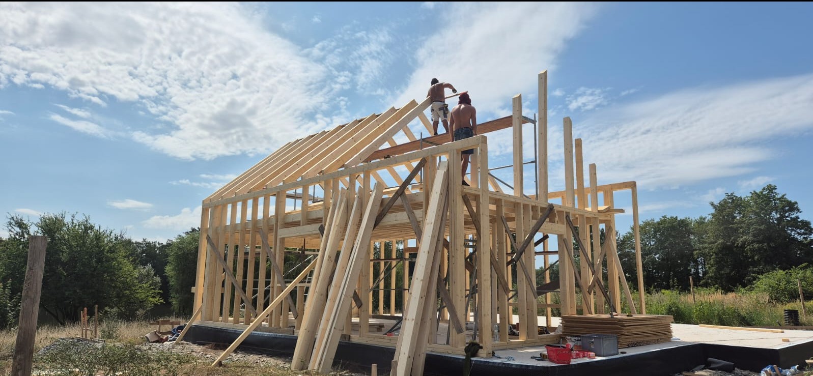 Konstrukcja szkieletowa domu w trakcie montażu, widoczni dwaj pracownicy na dachu, błękitne niebo z chmurami w tle.