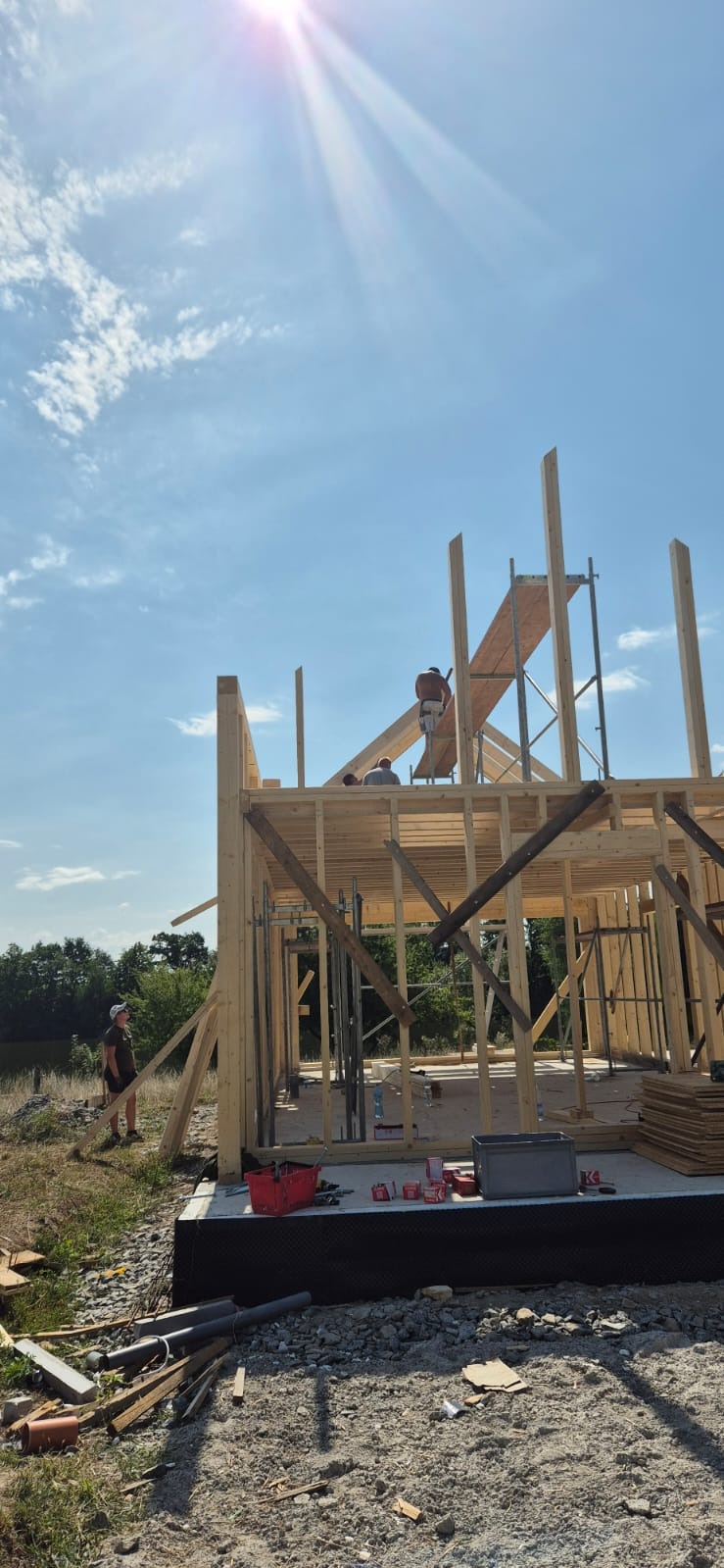 Wznoszenie konstrukcji drewnianej domu w słoneczny dzień, widoczni dwaj pracownicy na rusztowaniu i trzeci na ziemi, rozrzucone materiały budowlane w otoczeniu.