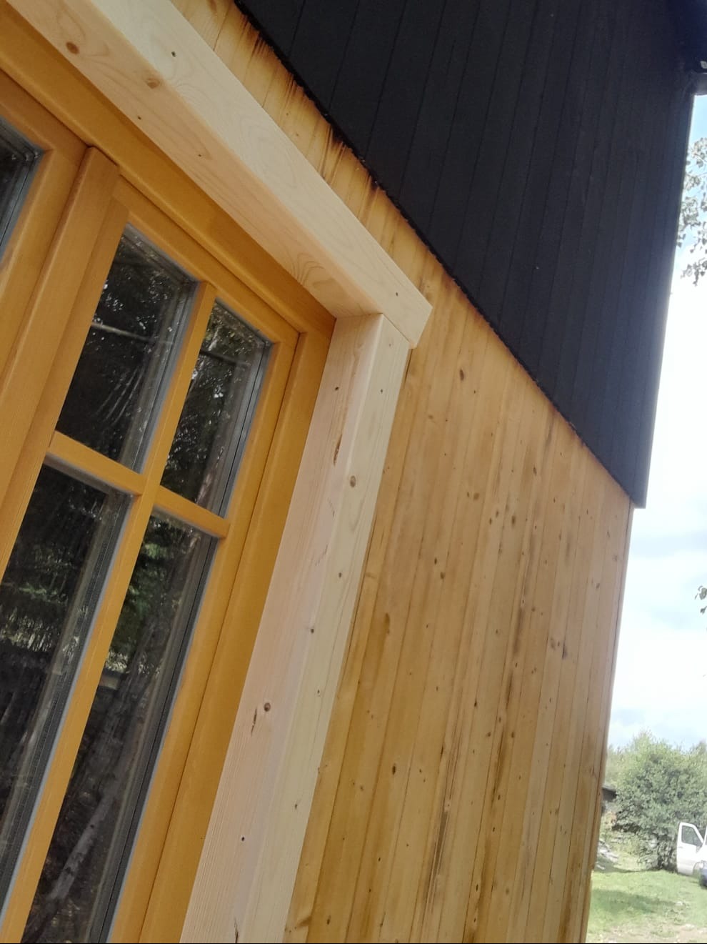 Fragment drewnianego domu z oknem w trakcie budowy, widoczne deski elewacyjne i surowe wykończenie okna.