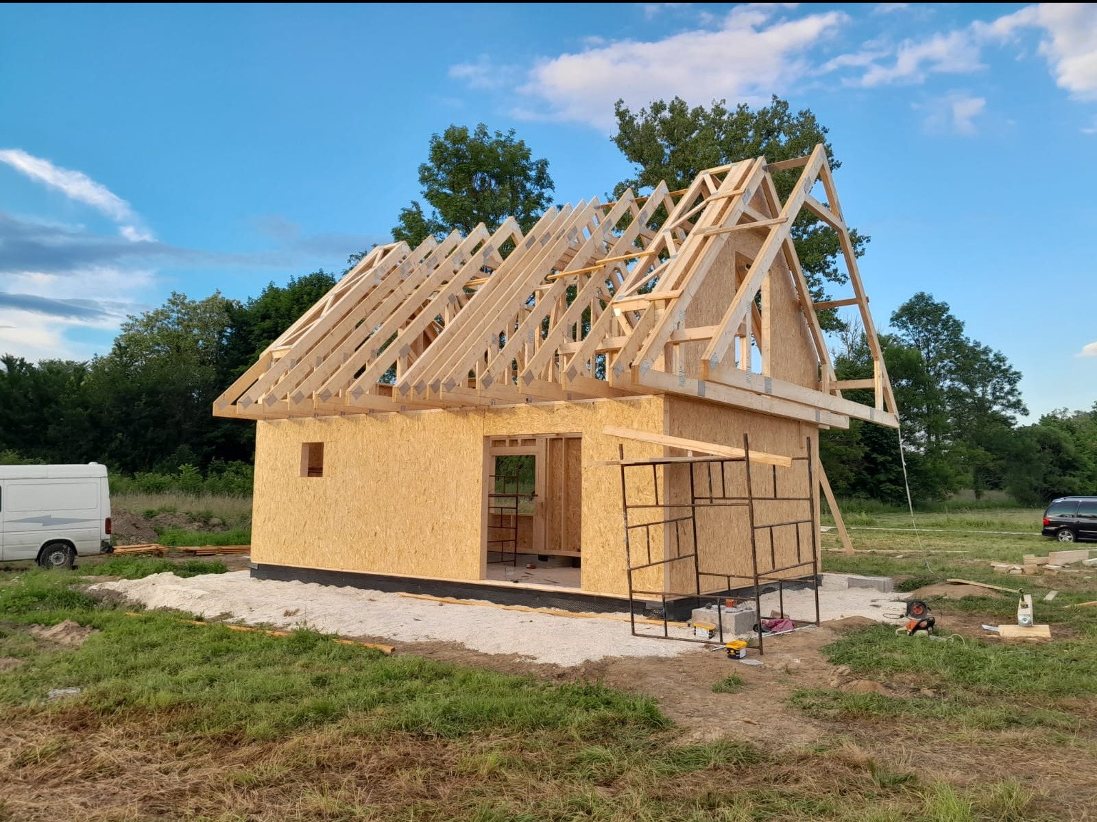 Wznoszenie konstrukcji drewnianego domu z płyt OSB, widoczne rusztowanie i elementy więźby dachowej na tle zieleni i błękitnego nieba.
