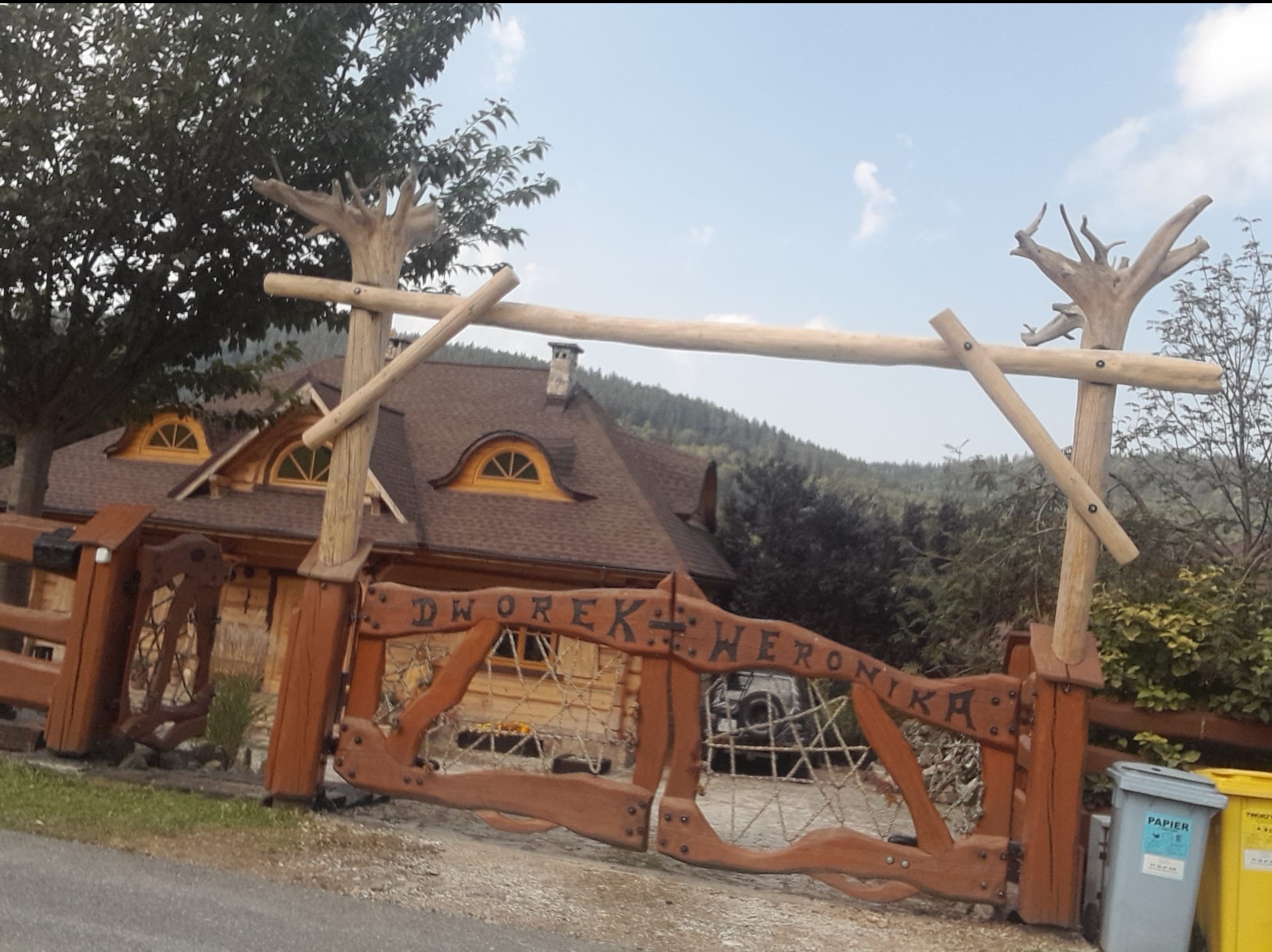 Drewniany dom z brązowym dachem i oknami w kształcie półksiężyca, widoczny zza ozdobnej bramy z napisem 'Dworek Weronika', zwieńczonej drewnianymi rzeźbami imitującymi drzewa.