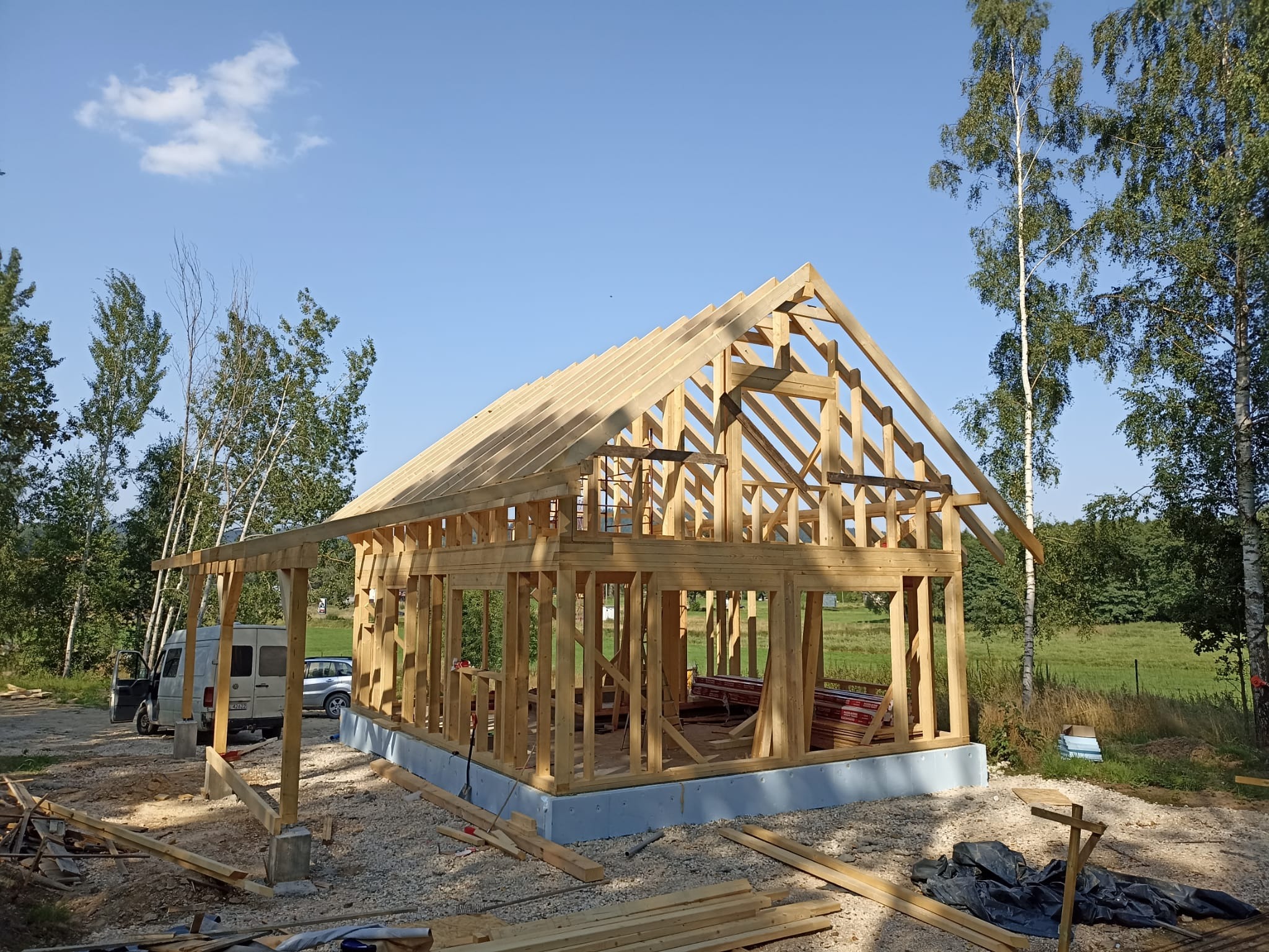 Surowa konstrukcja domu szkieletowego z drewnianych belek na fundamencie, w tle zieleń drzew i błękitne niebo. Widoczny samochód dostawczy i materiały budowlane.