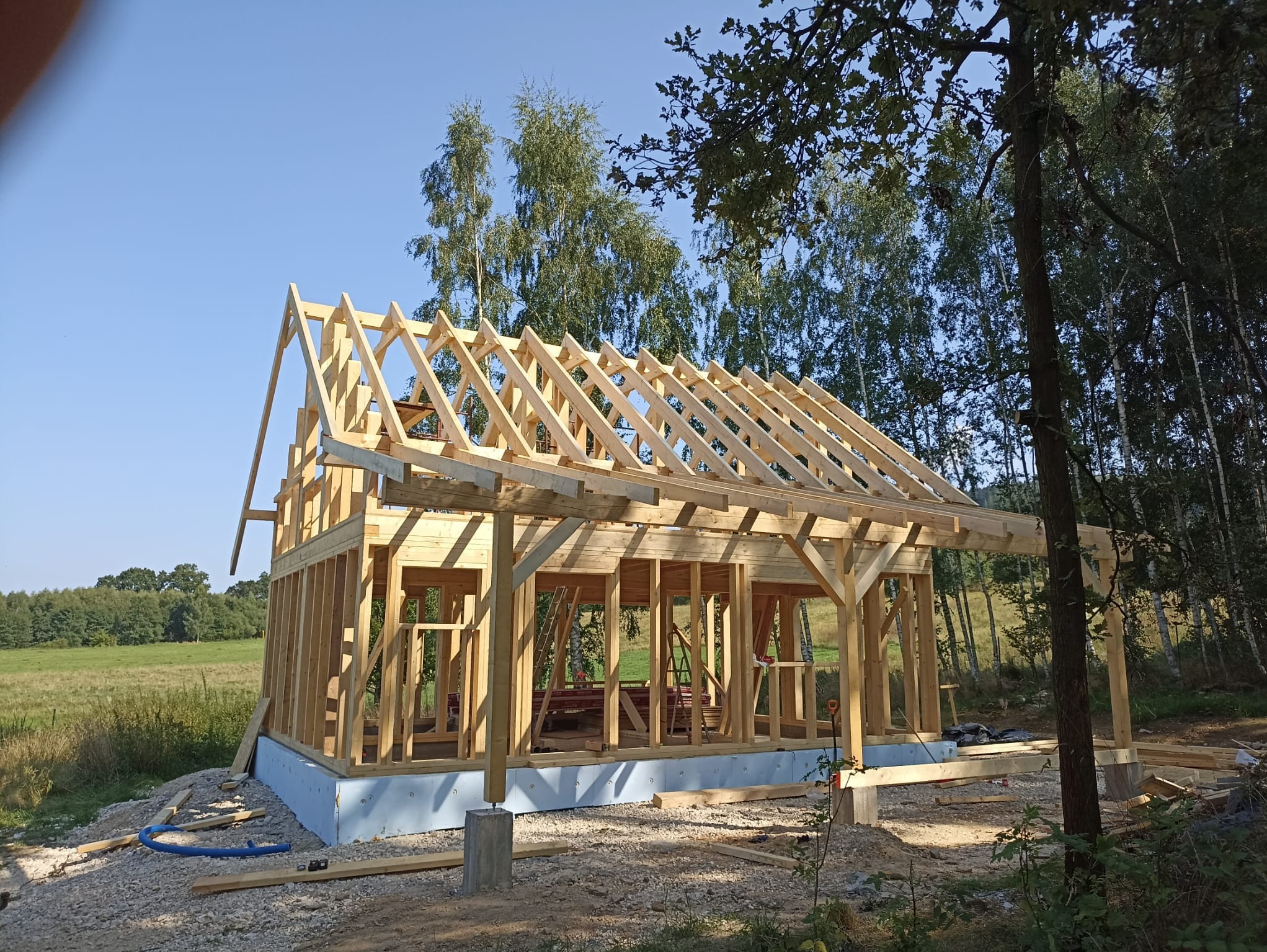 Surowa konstrukcja domu szkieletowego z drewnianych belek na fundamencie, widok z zewnątrz na tle zieleni i błękitnego nieba. Widoczne elementy złączne i narzędzia.