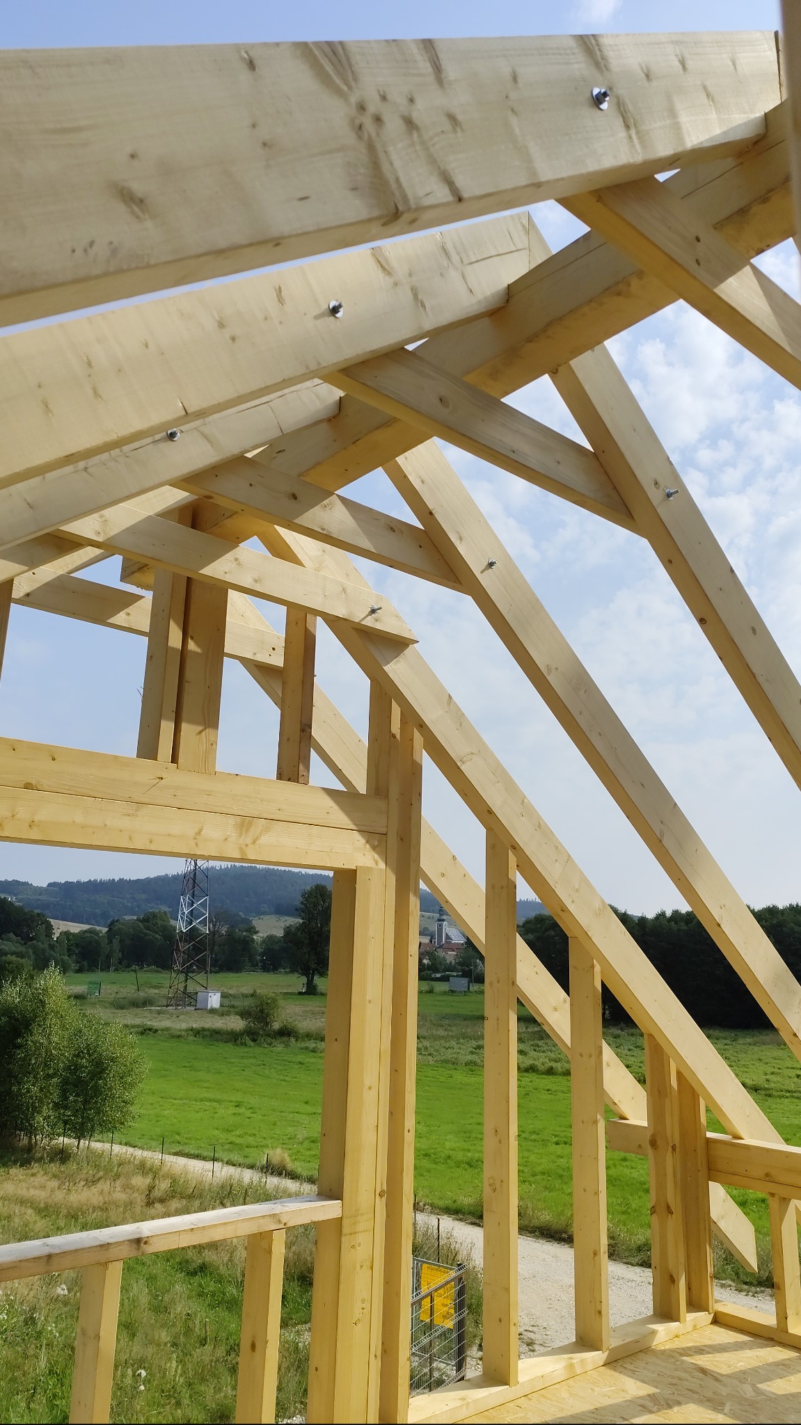 Surowa konstrukcja drewniana domu szkieletowego w trakcie budowy, widok z wnętrza na zielony krajobraz. Elementy złączone śrubami, jasne drewno.