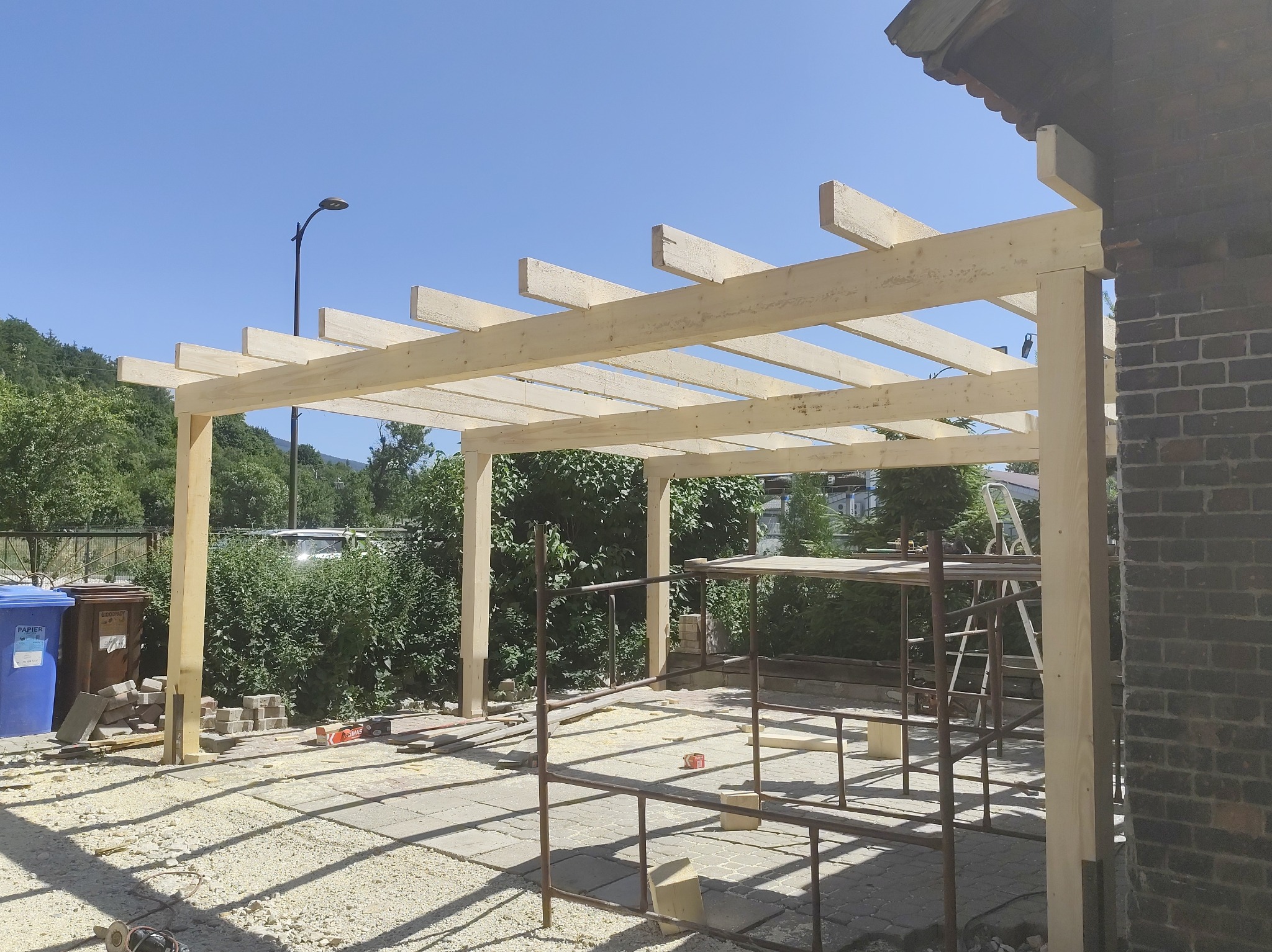 Drewniana konstrukcja z belek, prawdopodobnie zadaszenie tarasu, w trakcie budowy. Widoczne rusztowania i narzędzia. Tło: zieleń i błękitne niebo.