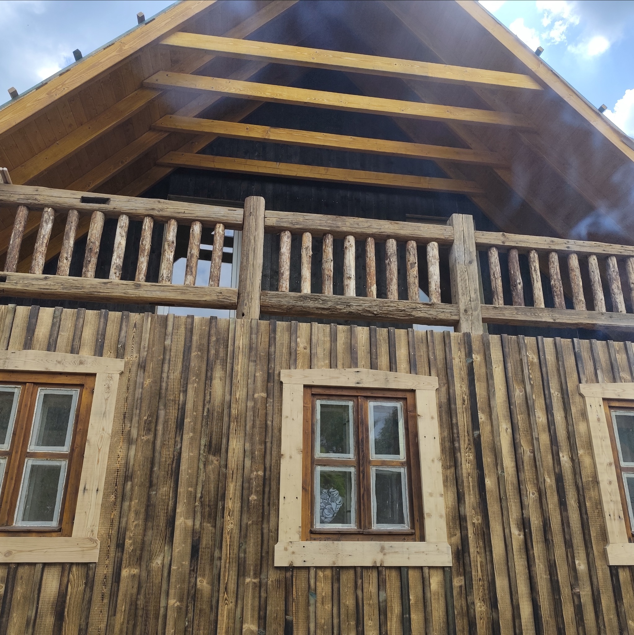 Drewniany dom z balkonem i oknami w rustykalnym stylu. Widoczne belki stropowe i deski elewacyjne z wyraźnym usłojeniem. Balustrada balkonu z okrąglaków.